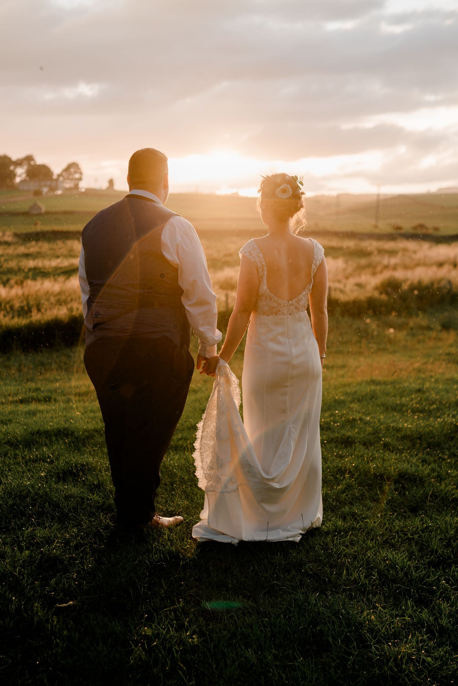 Bride and groom hold hands walking toward a breathtaking countryside sunset.