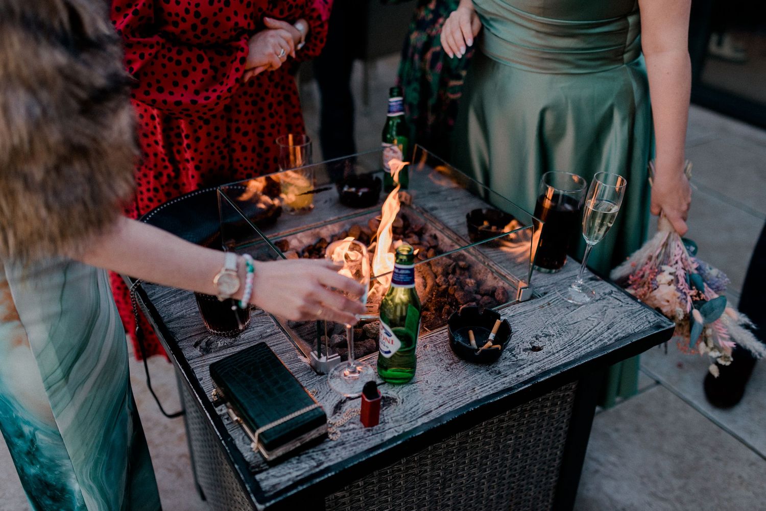 Wedding guests gathered around outdoor fire pit table with drinks and candles during evening wedding reception.