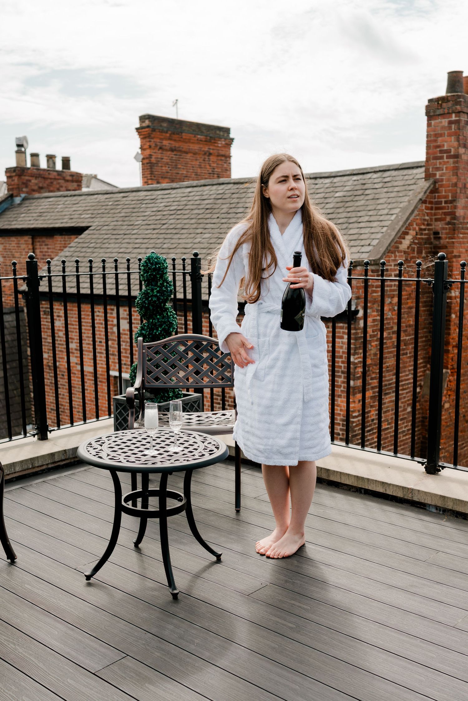 Woman in white bathrobe holding wine bottle on rooftop terrace with iron railings and brick buildings backdrop.