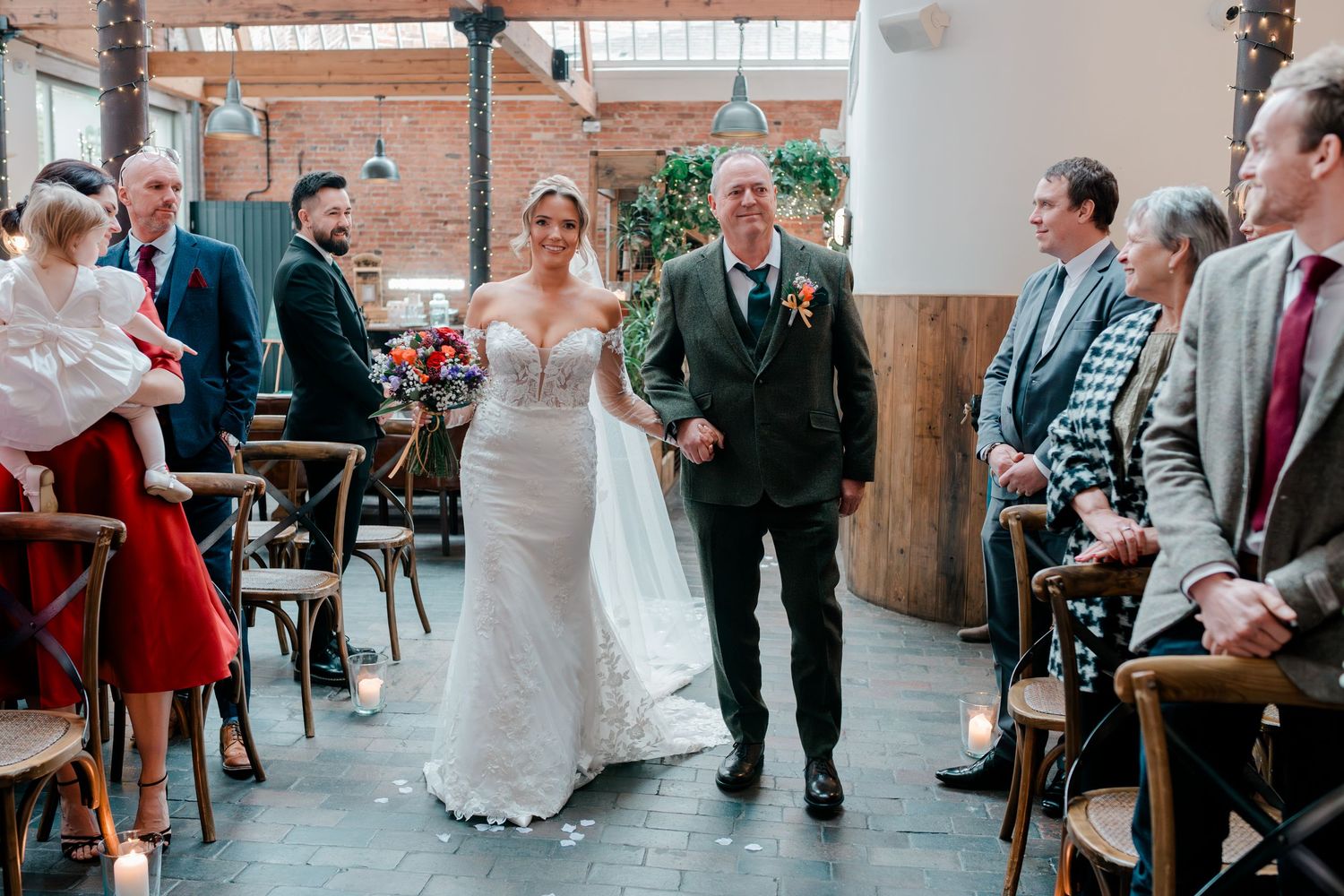 Bride walks down the aisle with escort in elegant lace wedding dress at rustic indoor ceremony.
