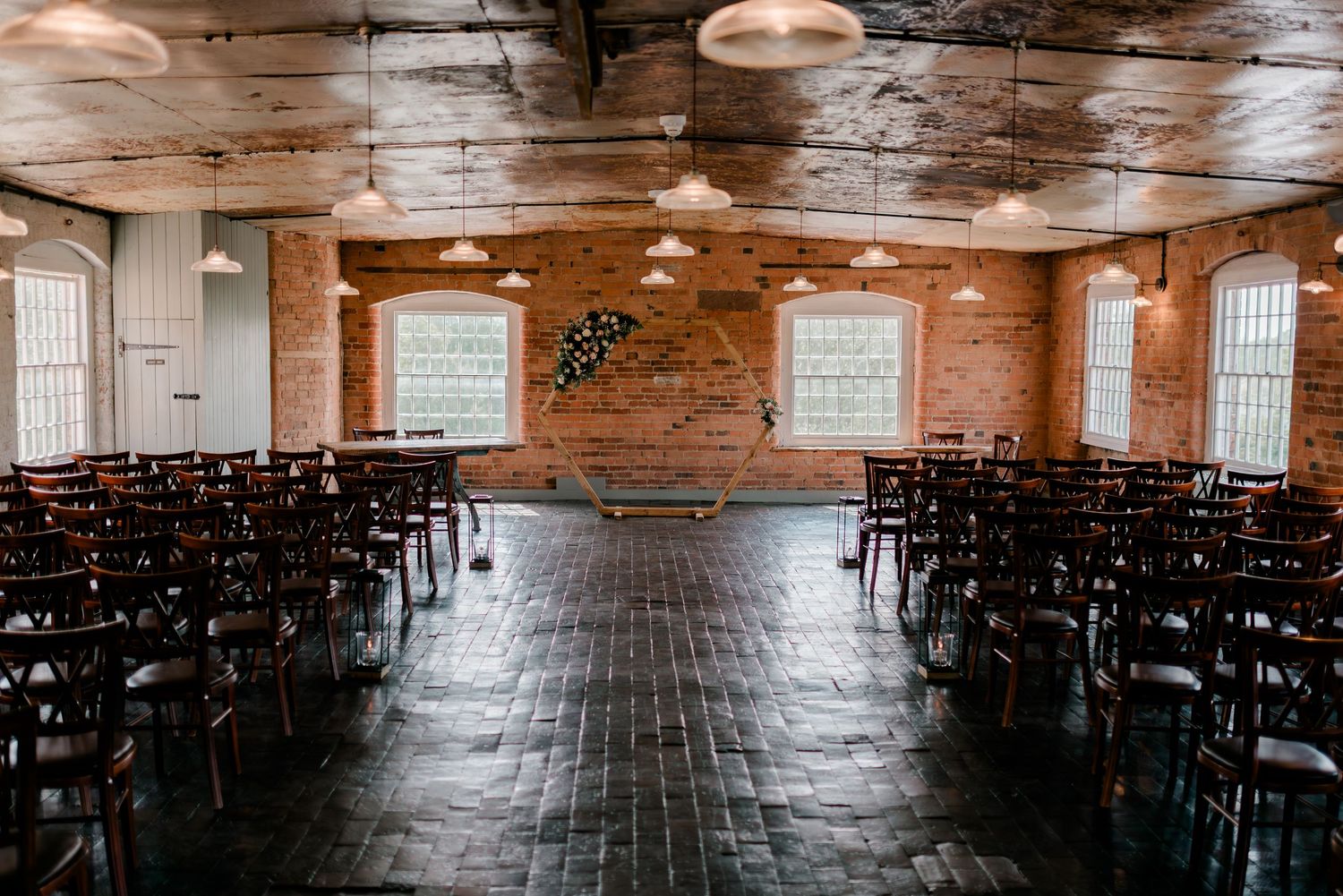 Industrial brick wedding ceremony space with dark cross-back chairs, pendant lights and floral arch backdrop.