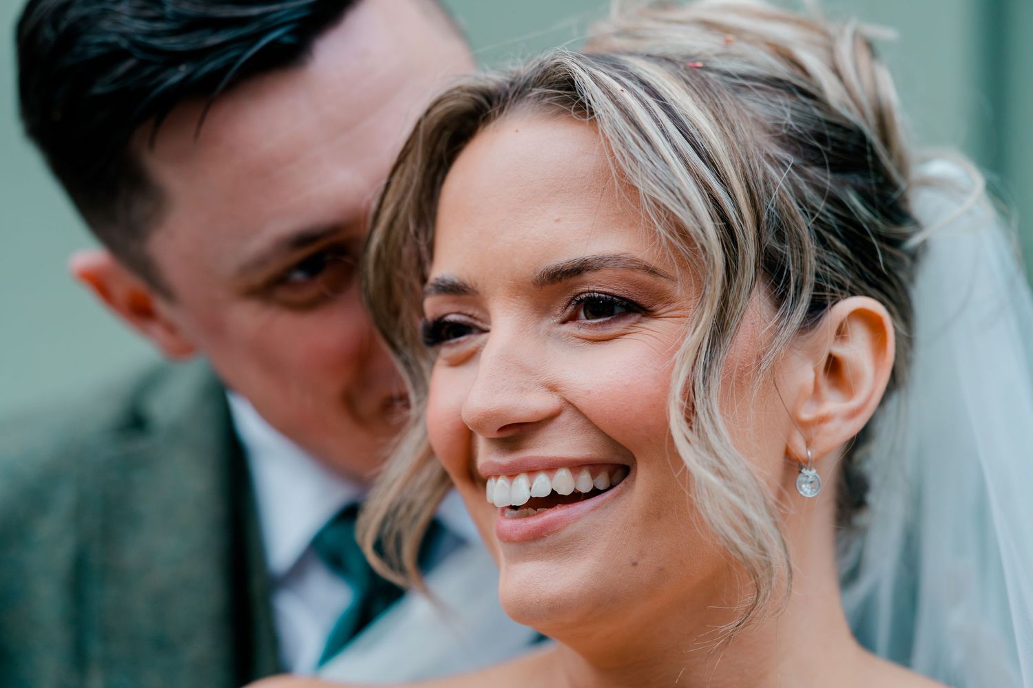Close-up of radiant bride smiling joyfully with groom in grey suit during their wedding day portrait.