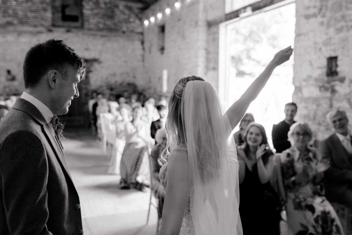 Black and white wedding photo showing celebration with bride raising hand in rustic stone venue.