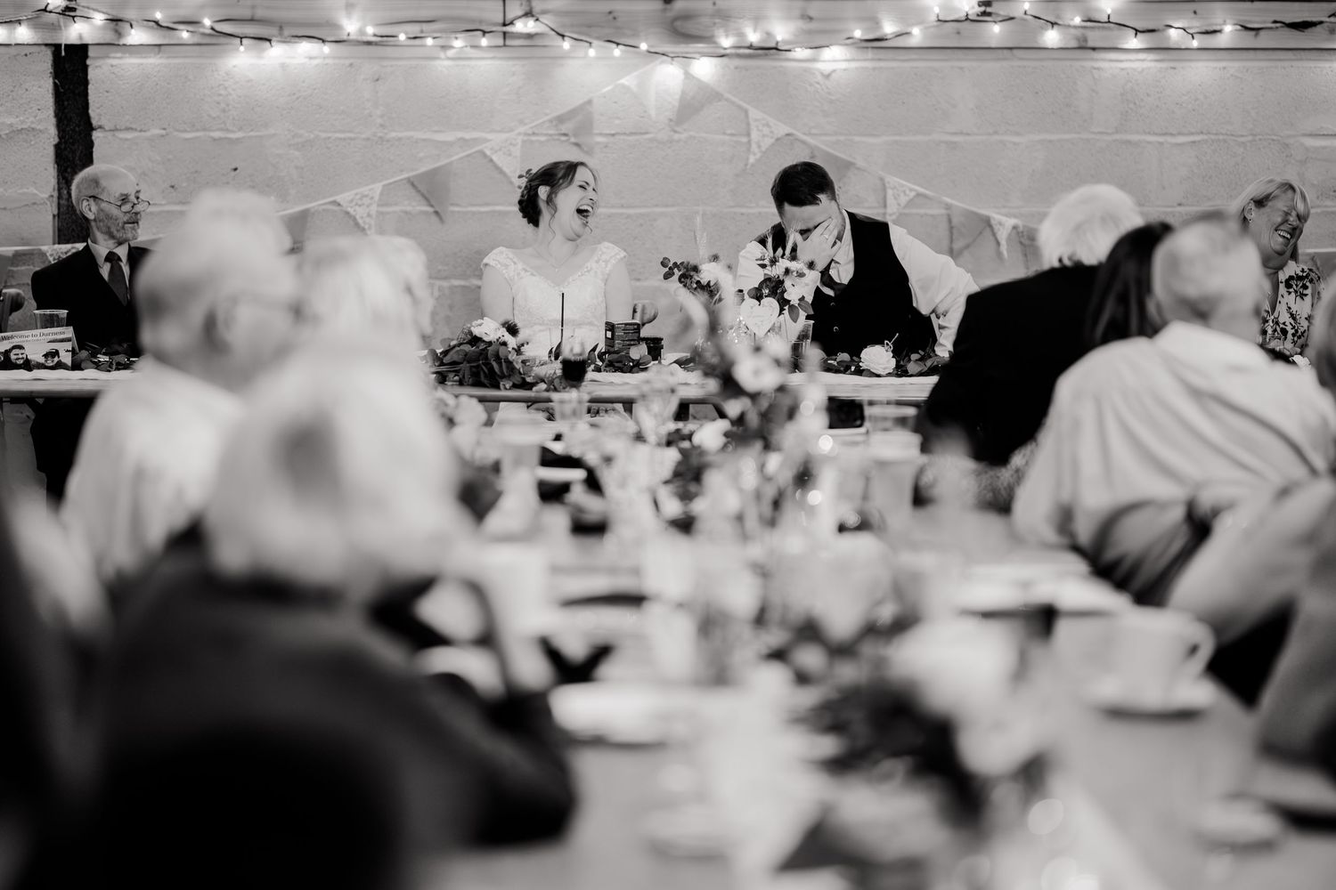 Black and white photo of bride and groom at head table during wedding reception speeches in barn venue.