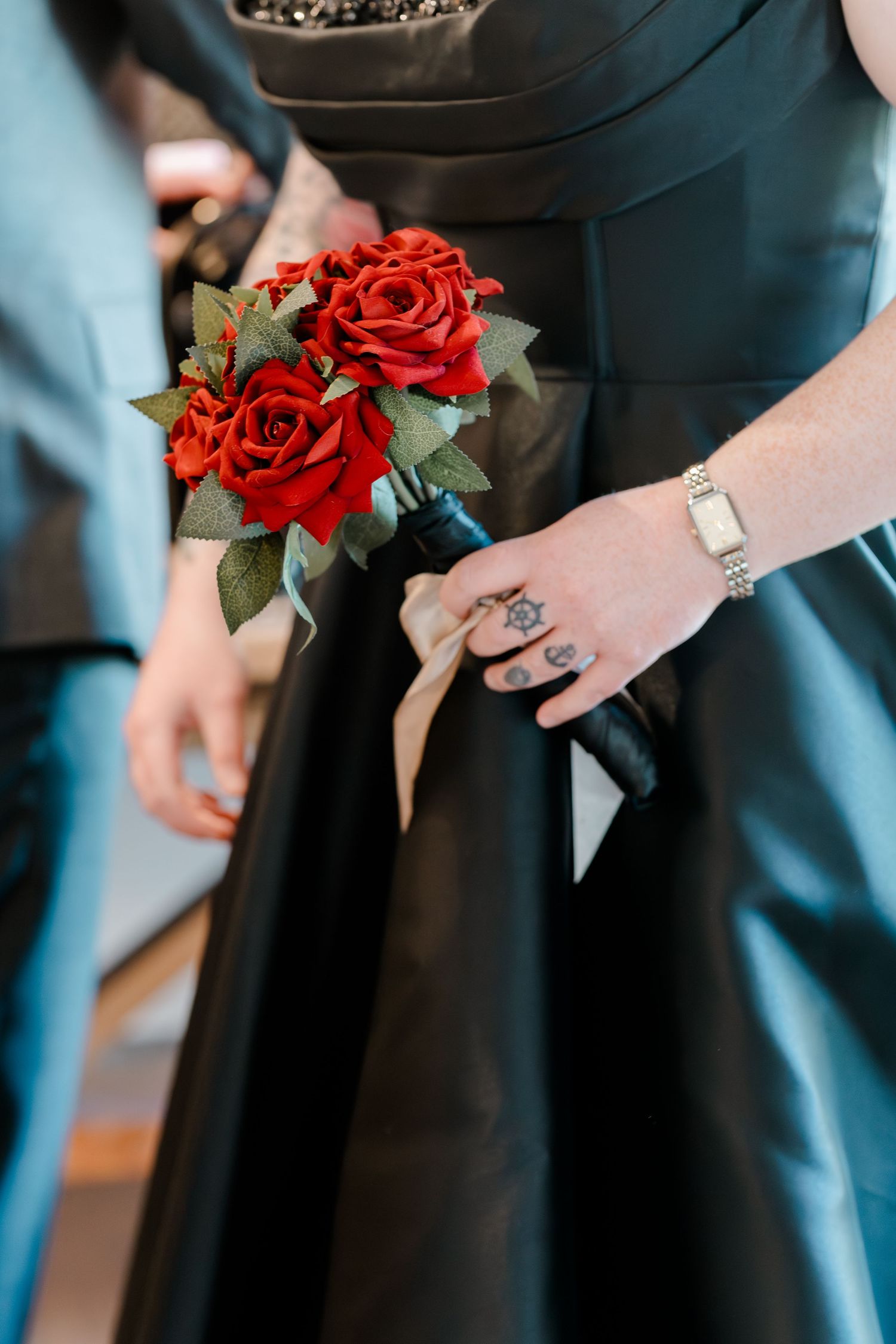 Tattooed hand holding red rose bouquet with black ribbon wrap, wearing silver watch and rings, against black satin gown.