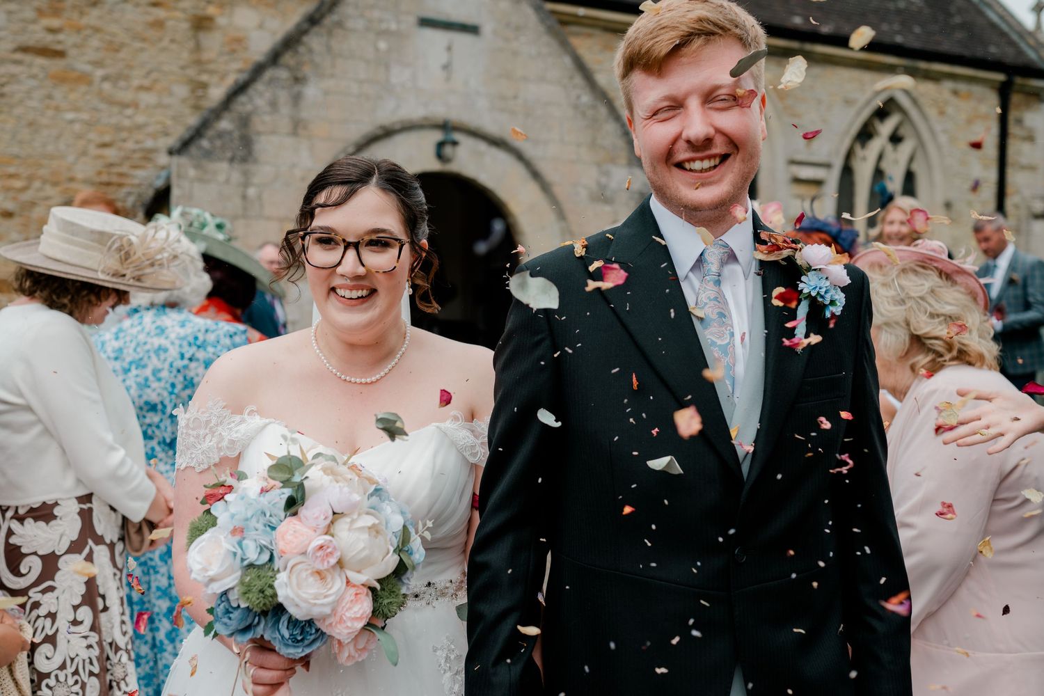 Happy newlyweds laughing as colorful confetti falls outside a stone church after their wedding ceremony.