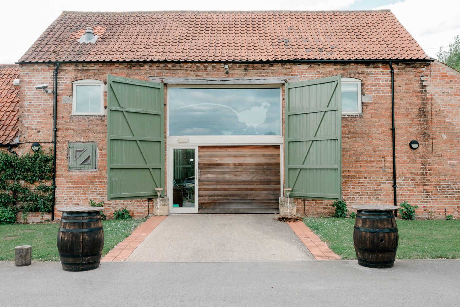 Rustic brick barn venue with open green wooden doors, wooden barrels, and a modern glass interior entrance.