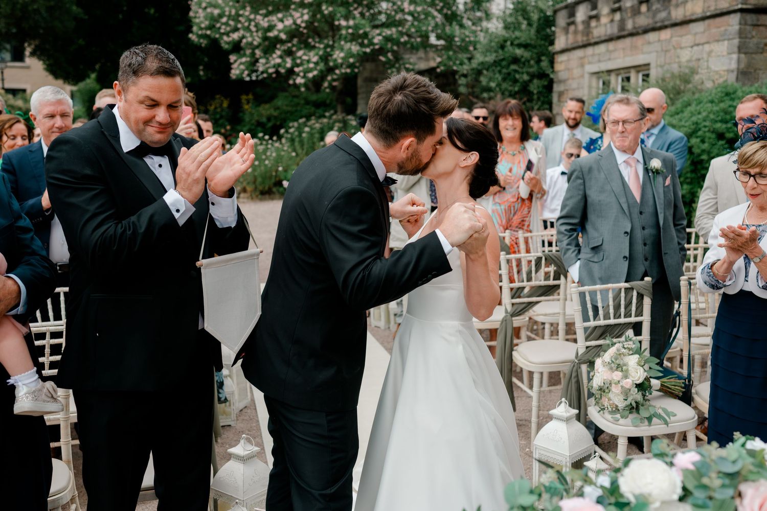 Bride and groom share a kiss during an outdoor wedding ceremony while smiling guests applaud beside white chairs and flowers.