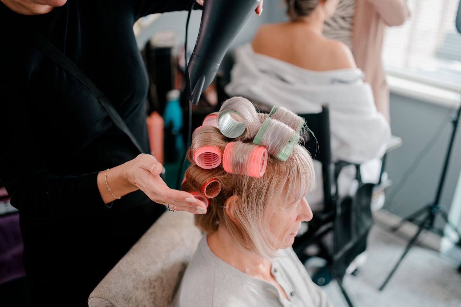 Hairstylist blow-drying hair with rollers for a wedding guest during bridal preparations.
