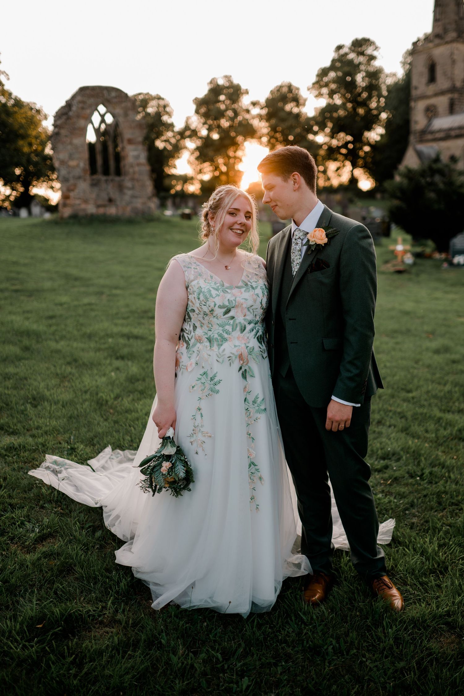 Bride in floral gown and groom pose at sunset near historic ruins.