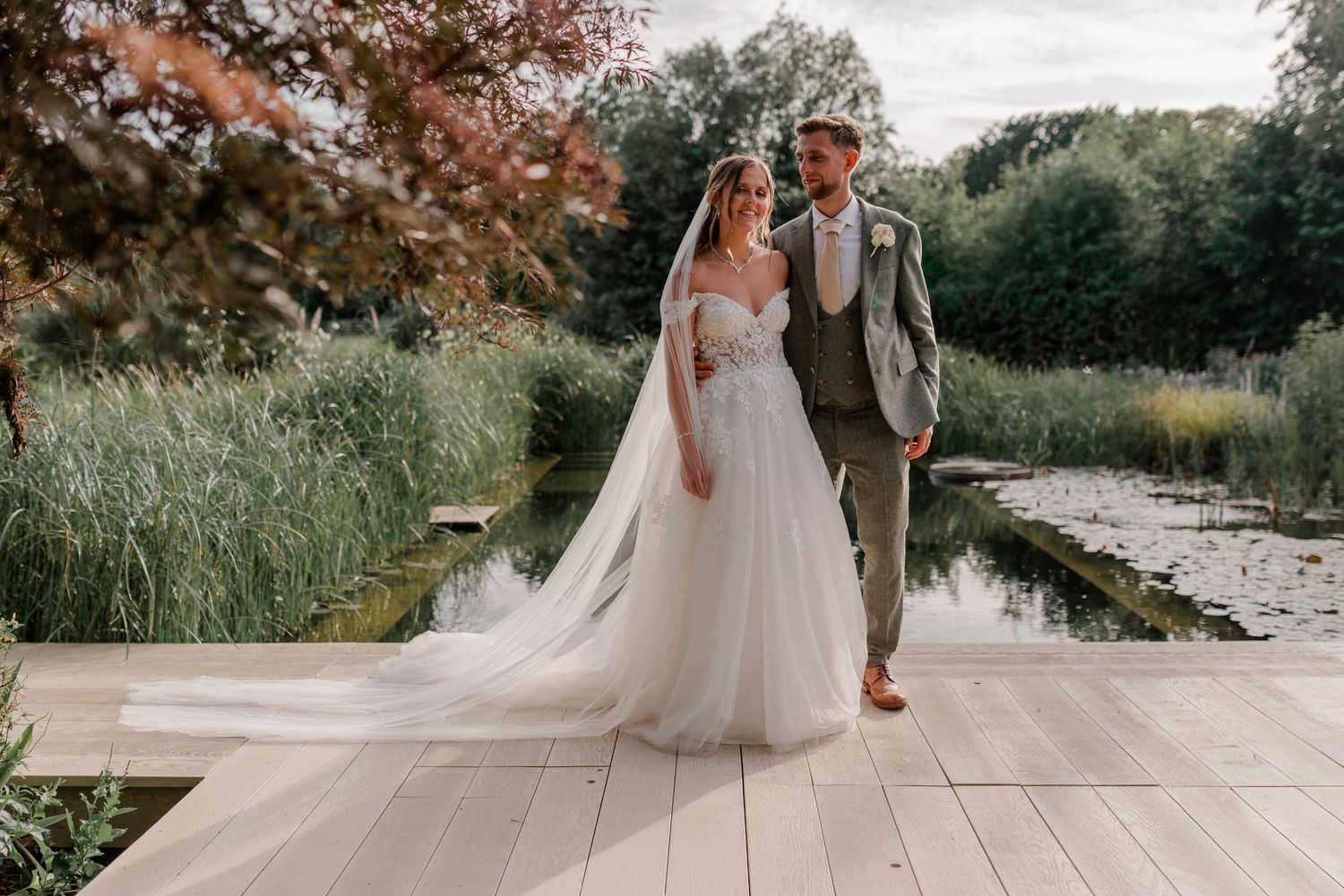 Wedding couple stands together on wooden deck overlooking pond with lush greenery and fall foliage in background.