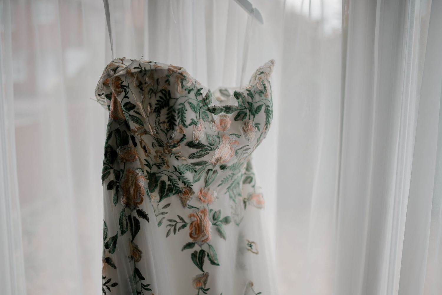 Delicate floral patterned wedding dress hanging against white curtains.