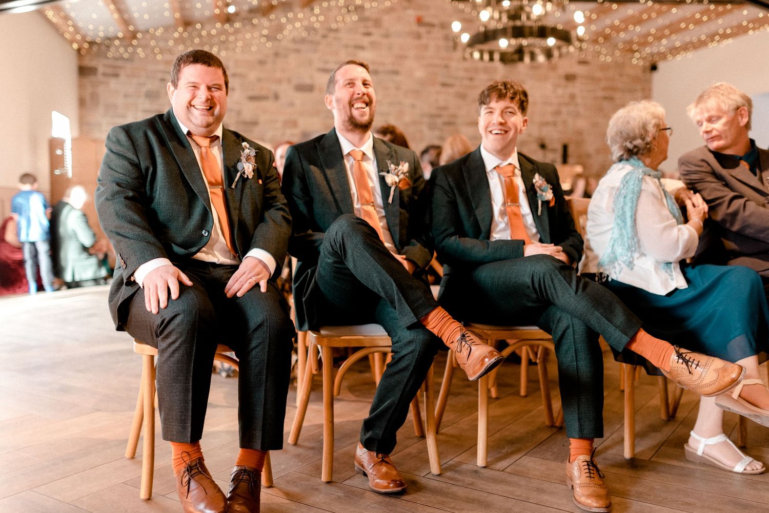 Wedding guests in matching orange ties seated on wooden chairs at reception venue.