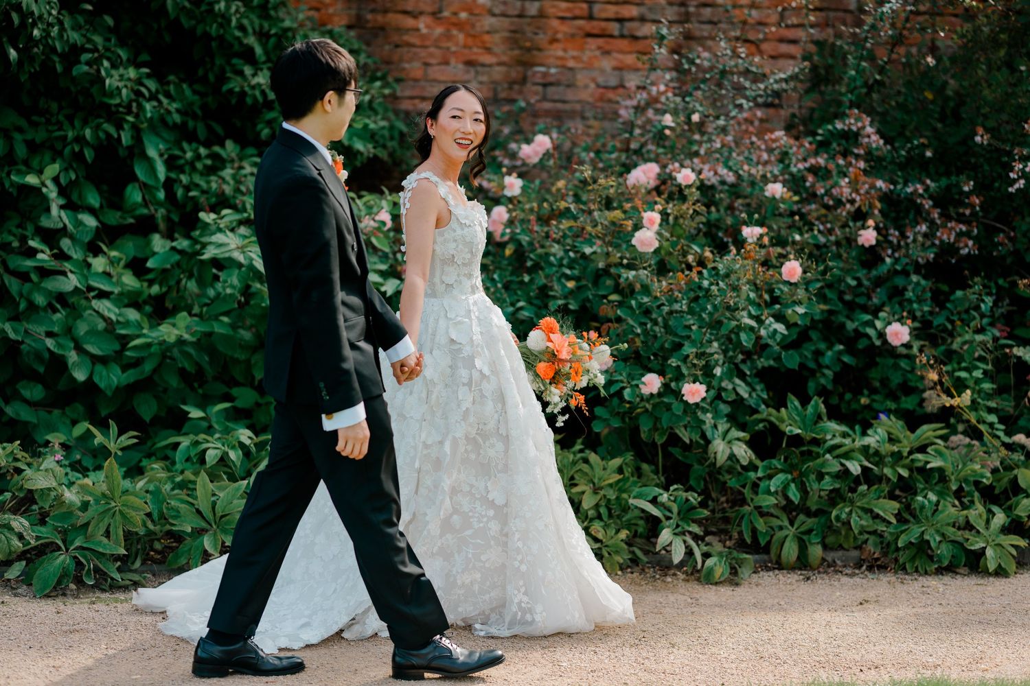 A bride in a floral gown and groom in a tuxedo walk together in a lush garden with blooming roses.