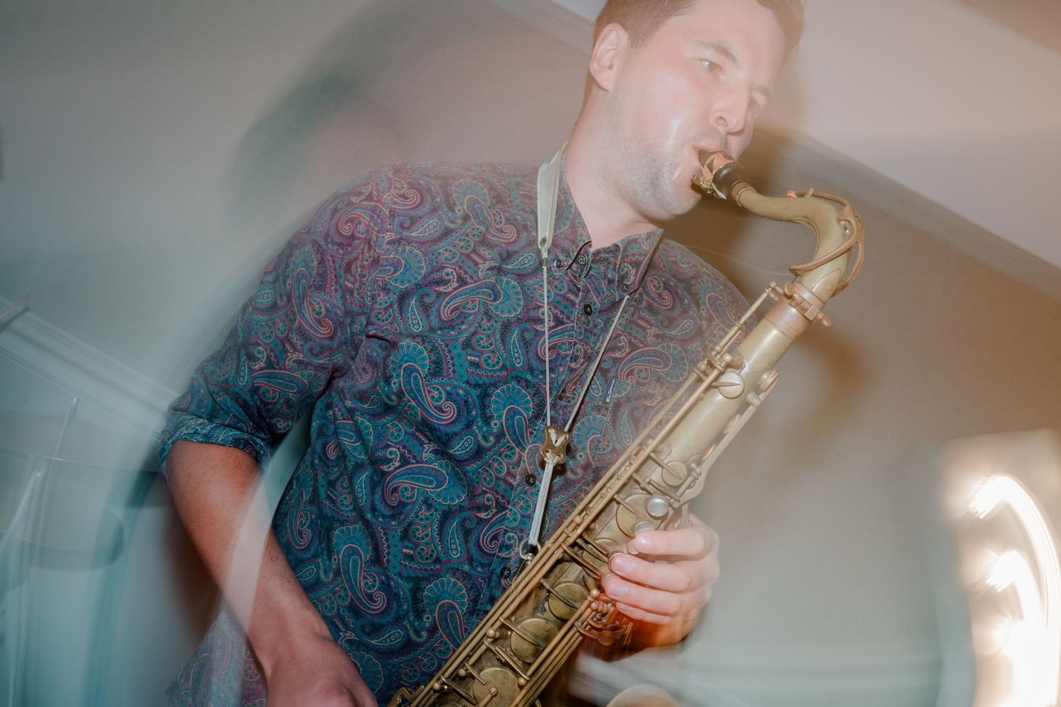 A saxophonist in a paisley patterned shirt plays a tenor saxophone, captured with energetic motion blur during a live performance.