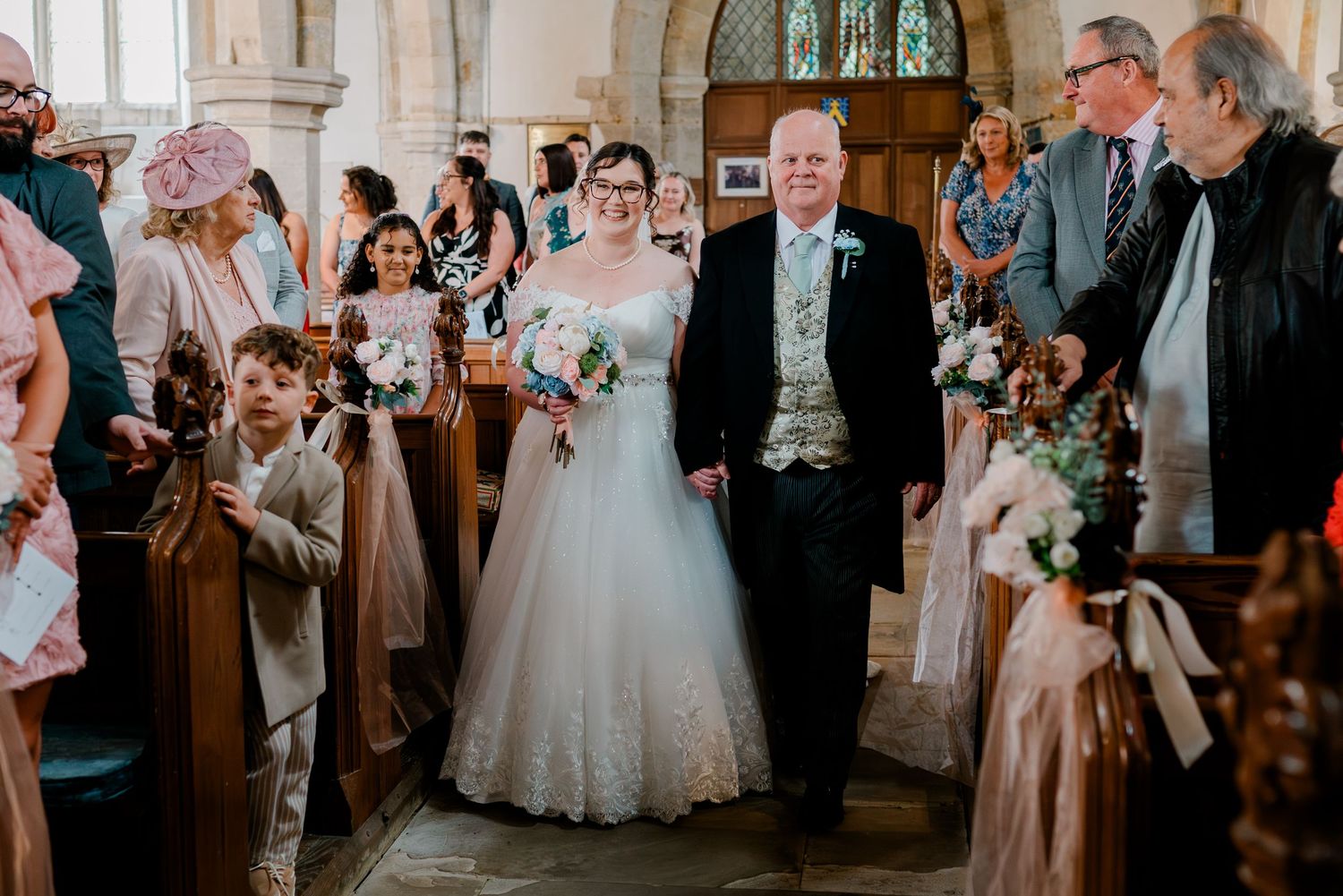 Bride walks down church aisle with father during wedding ceremony, guests seated in wooden pews.