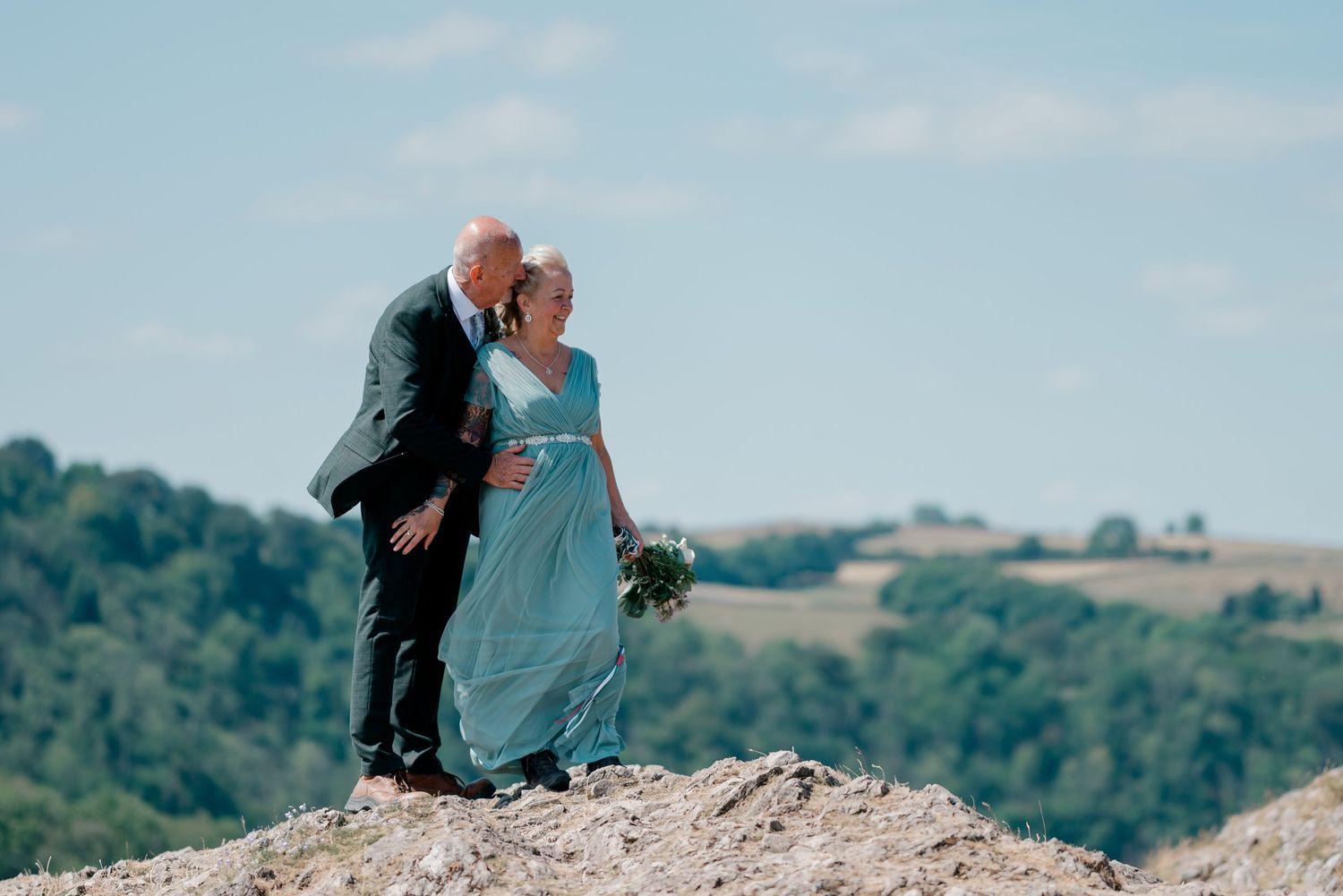 A couple in wedding attire shares a kiss on a rocky hilltop with a scenic green landscape under a blue sky.