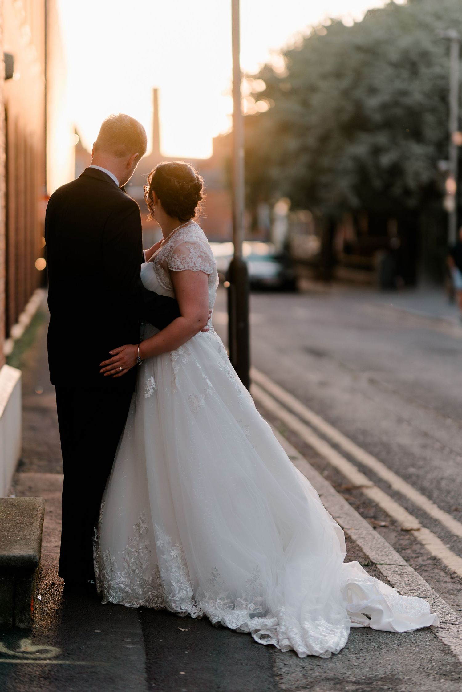 Bride and groom embrace on a sunlit city street, her lace ball gown trailing as golden hour glows behind them.