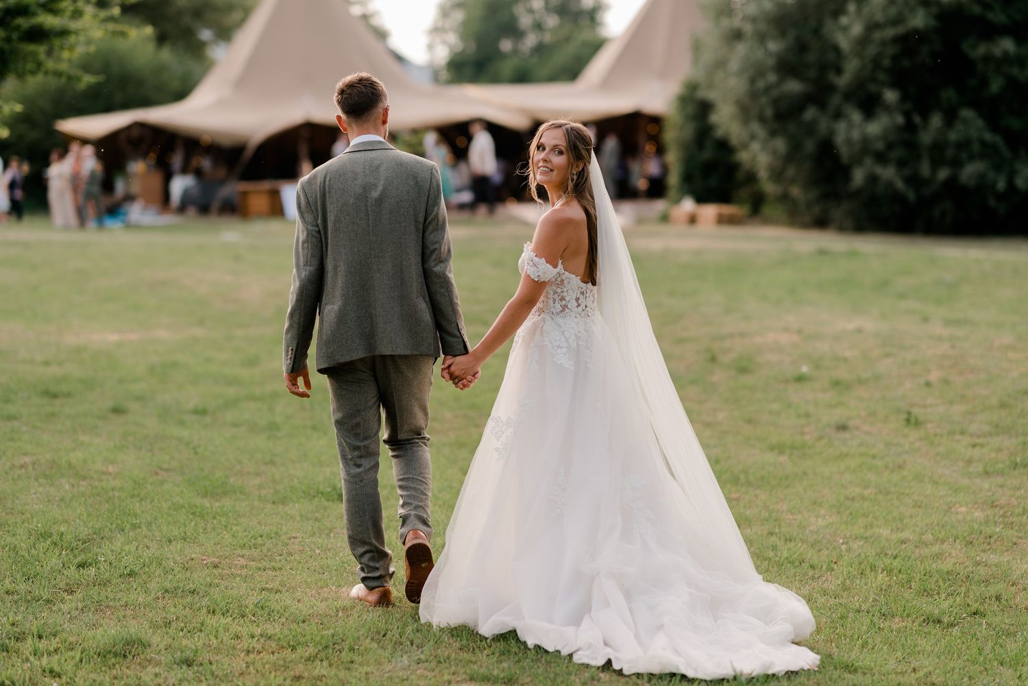 A couple in wedding attire holds hands while walking across a grass field at a garden ceremony.