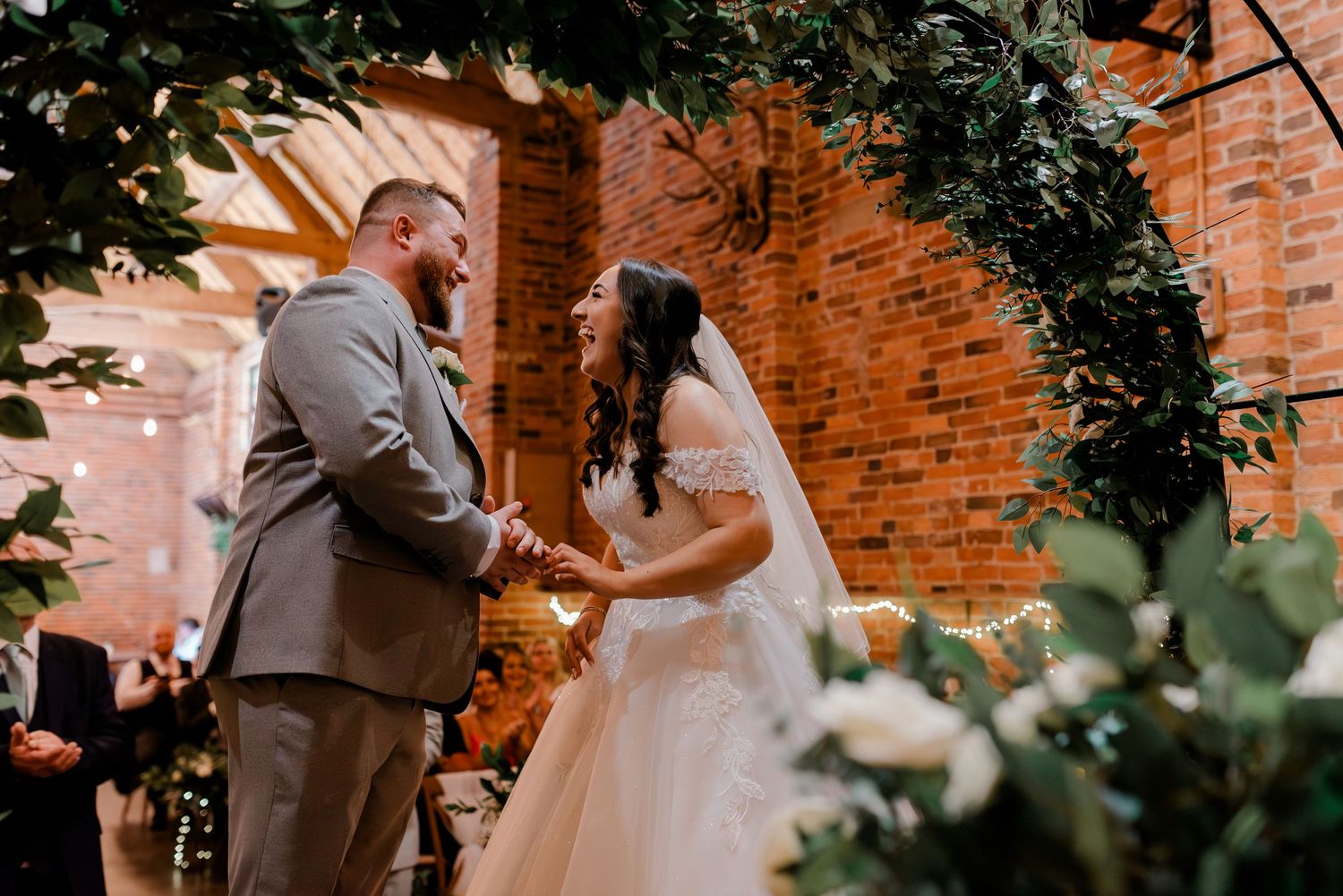 An intimate wedding ceremony under a lush greenery arch in a rustic brick venue.