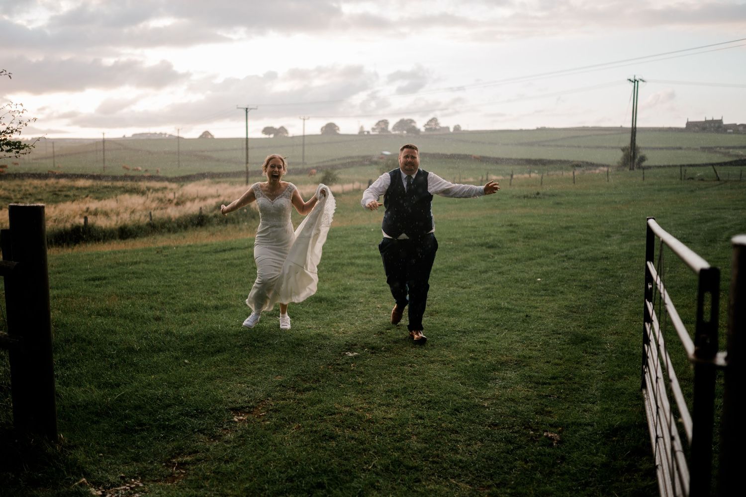 Wedding couple walks through scenic countryside field at sunset while photographer captures romantic moments.