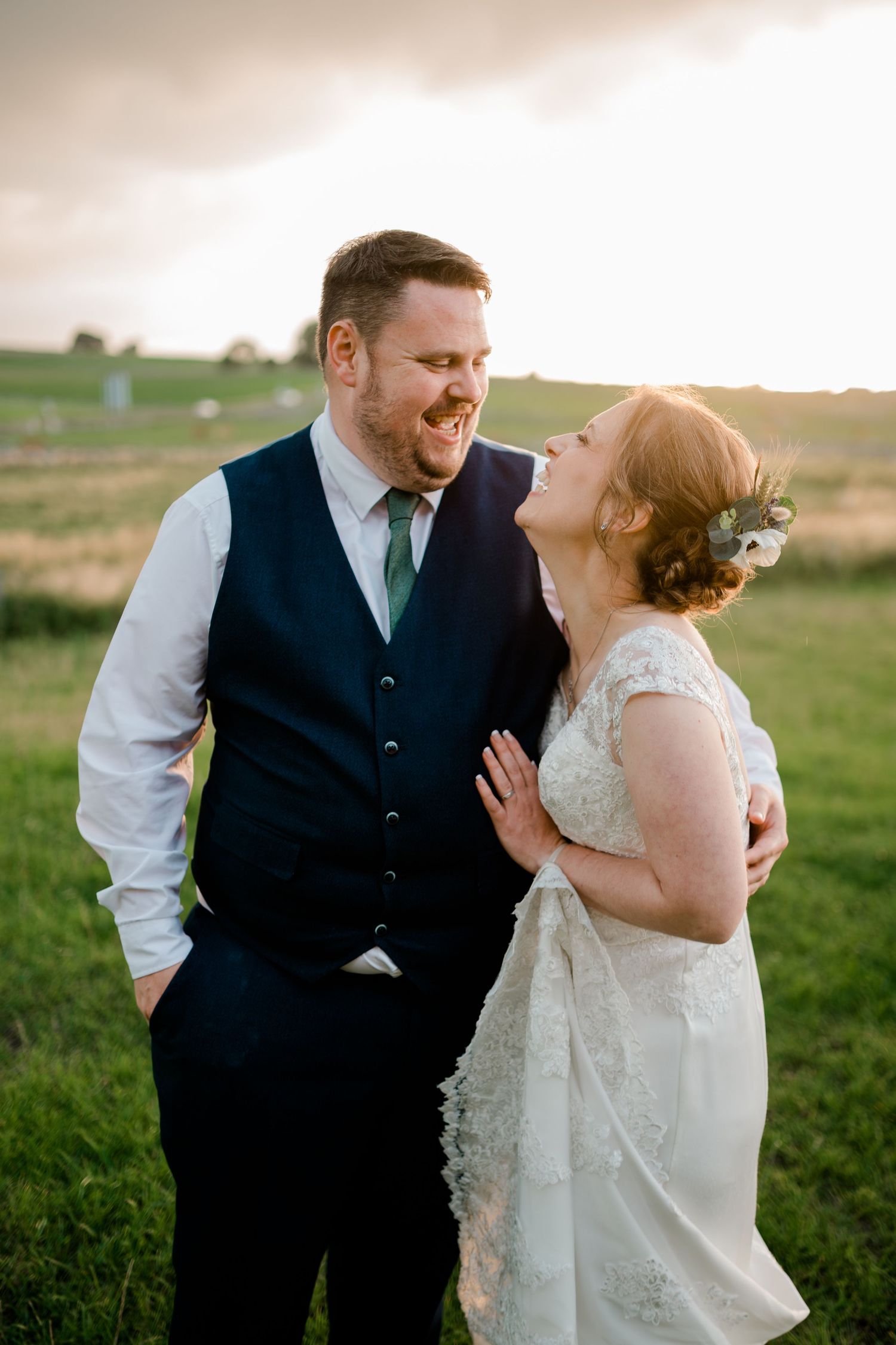 Laughing bride and groom sharing a joyful moment in a green field at golden hour during their wedding.