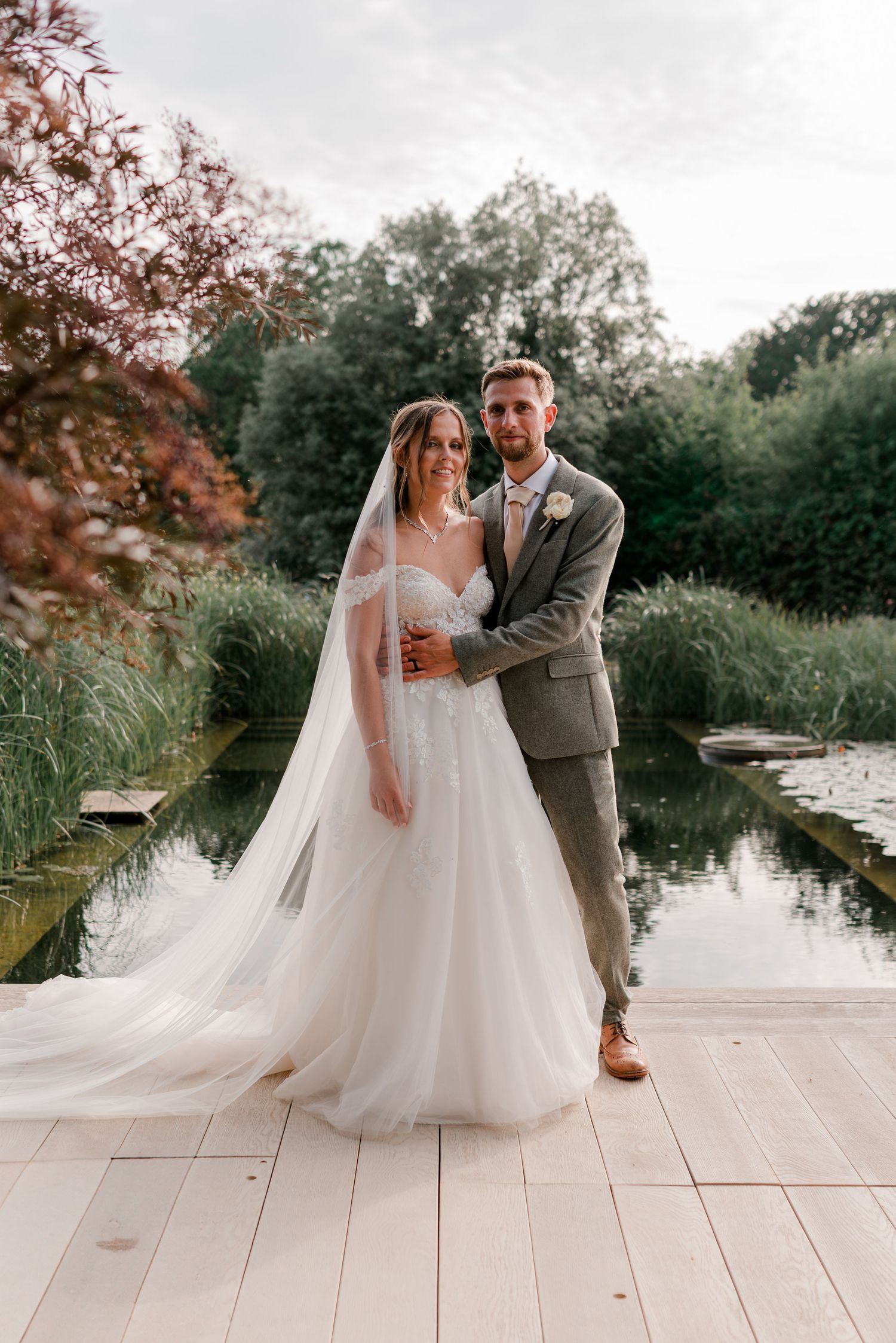 Bride and groom posing together on a wooden deck beside a serene pond surrounded by lush greenery.