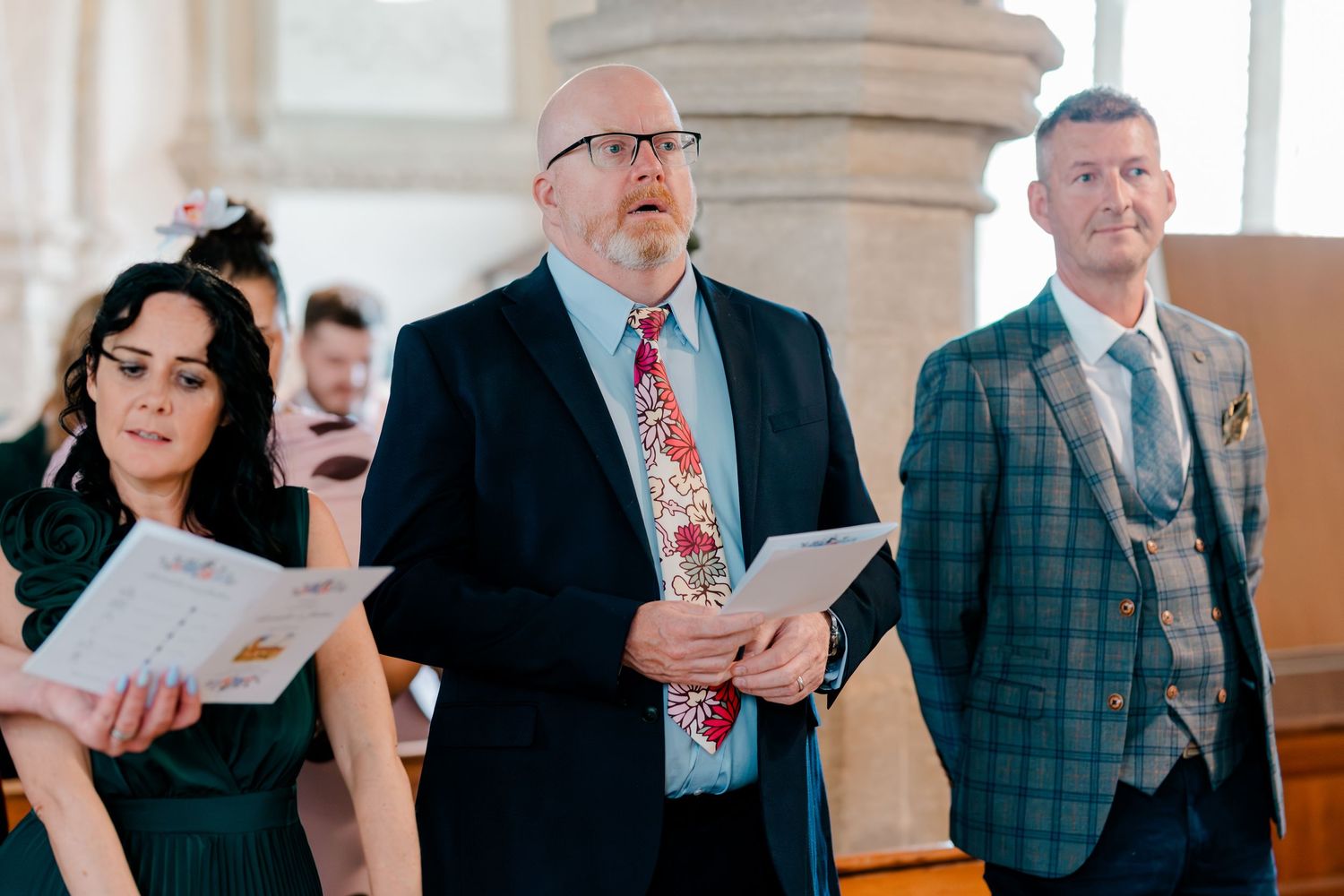 Wedding guests singing hymns in a church ceremony, holding order of service booklets.