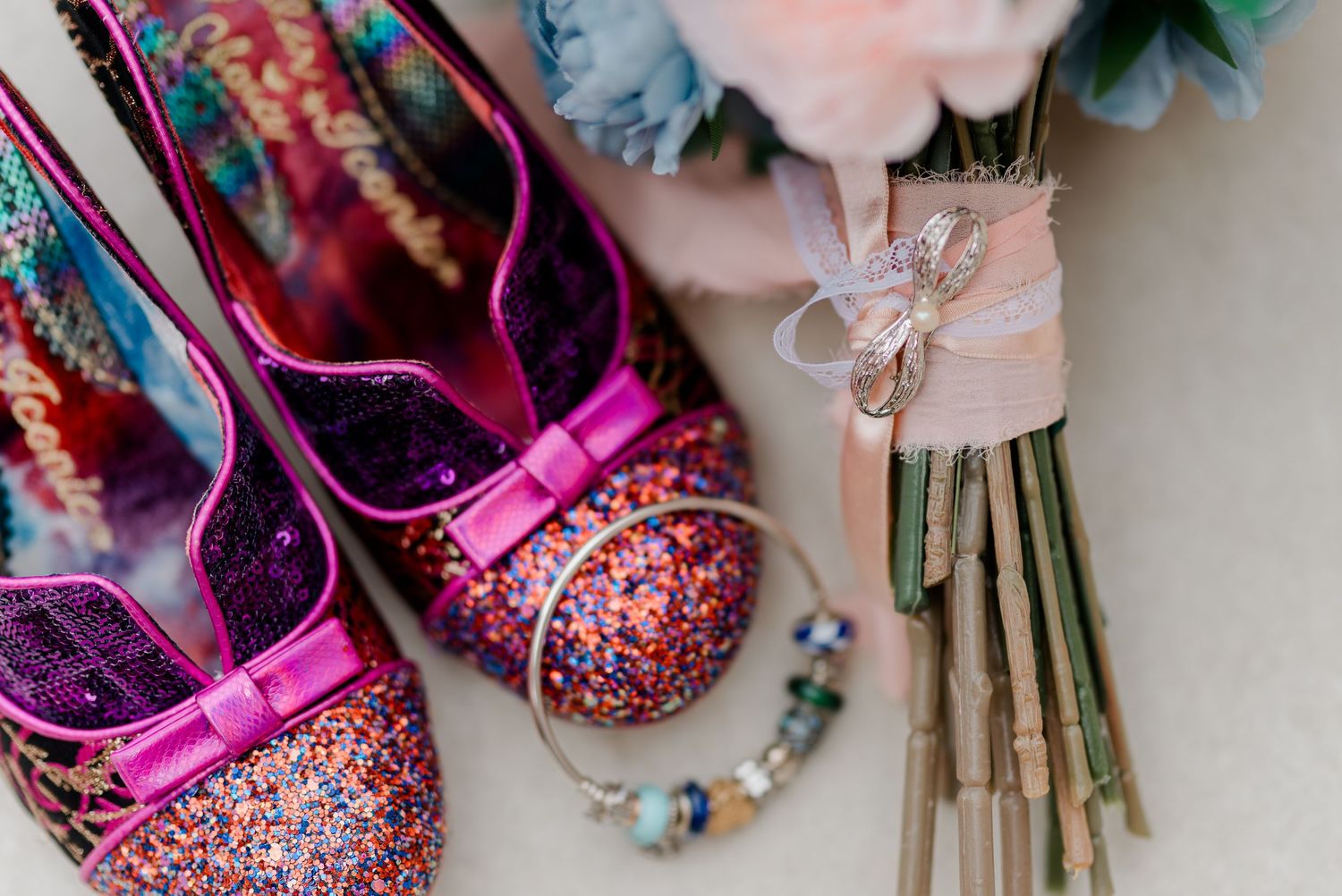 Sparkling pink glitter heels with bow detail beside a bridal bouquet and colorful bracelet on a white surface.