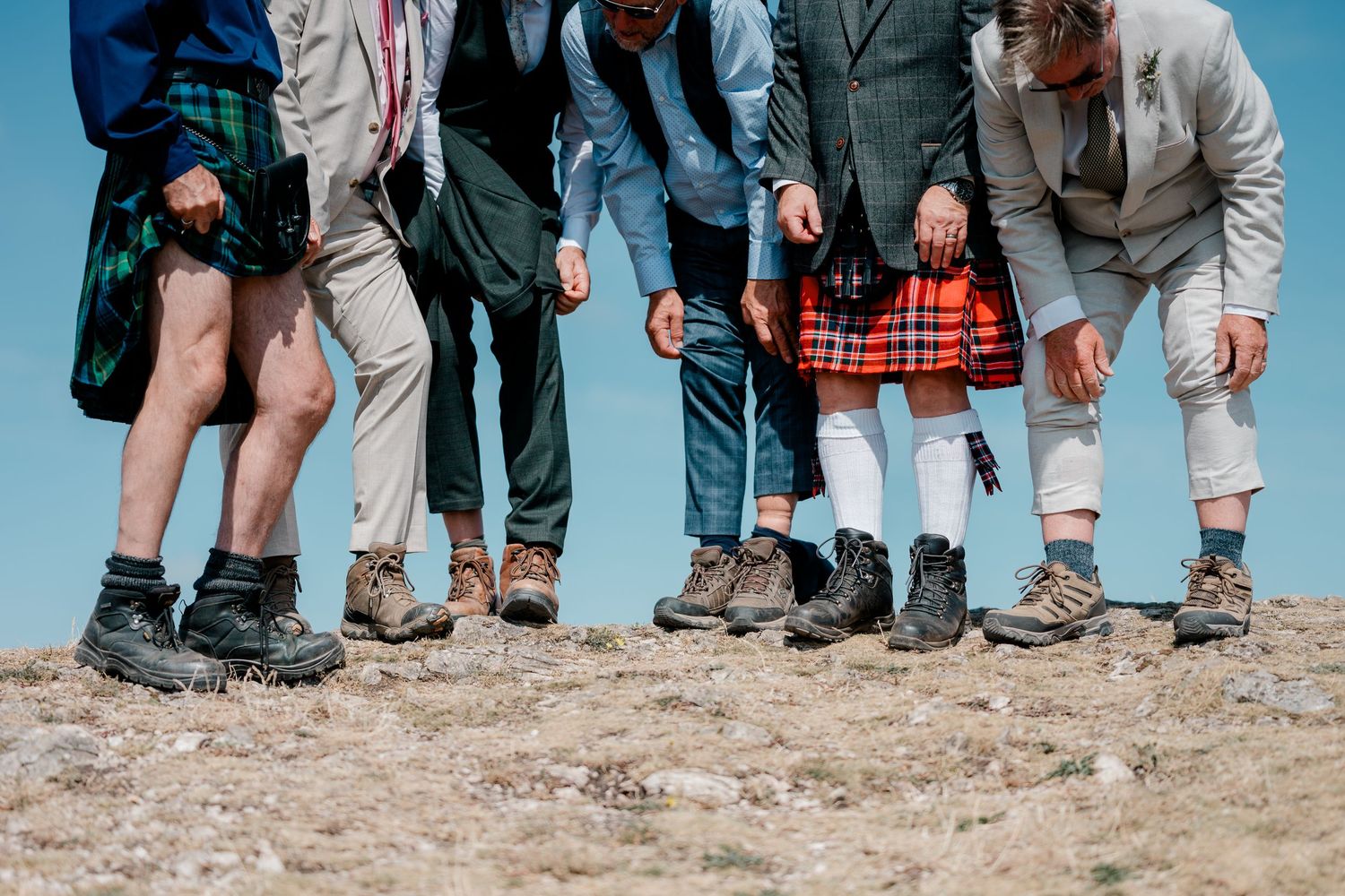 Wedding guests wearing kilts and hiking boots stand together on rocky ground.
