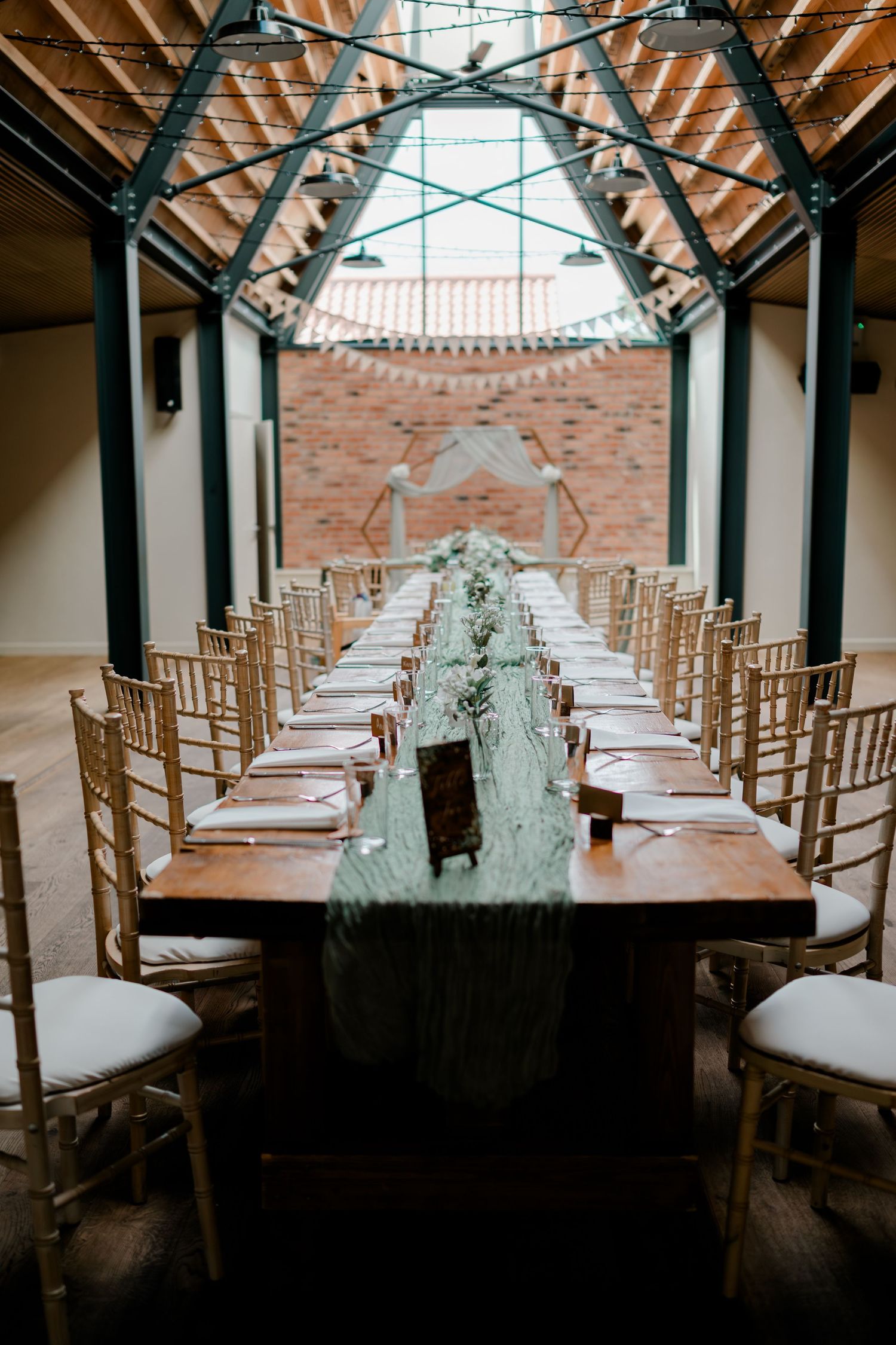 Elegant wedding reception setup with long wooden table, sage green runner, gold chiavari chairs in a rustic greenhouse venue.