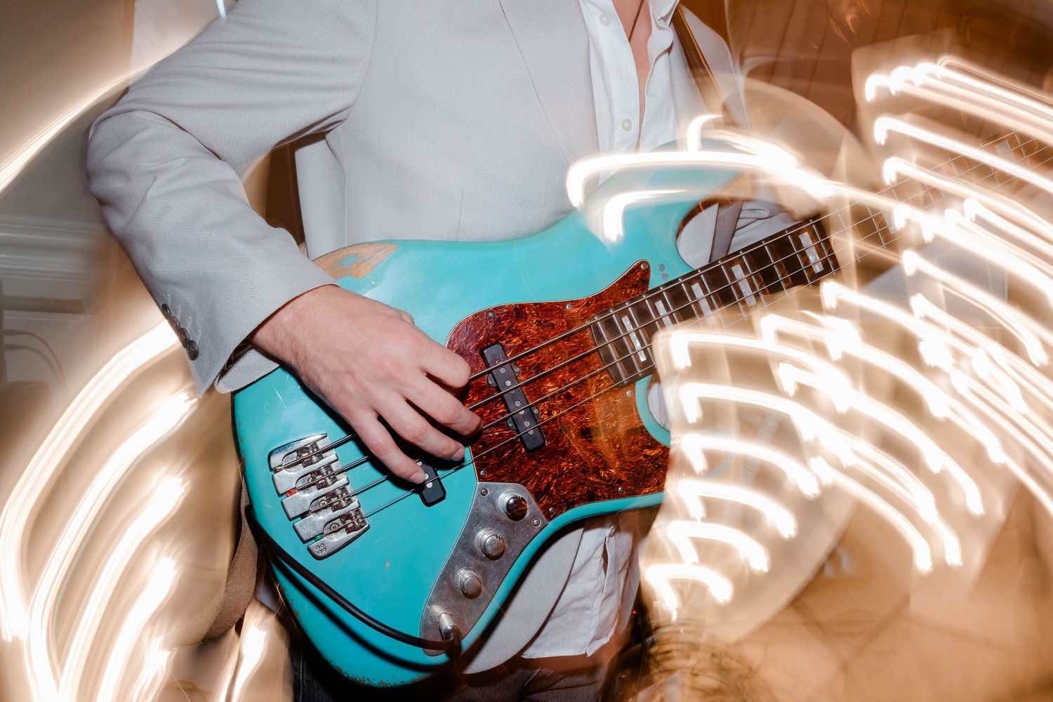 Musician in a light gray blazer playing a turquoise bass guitar with a tortoiseshell pickguard, surrounded by swirling light trails.