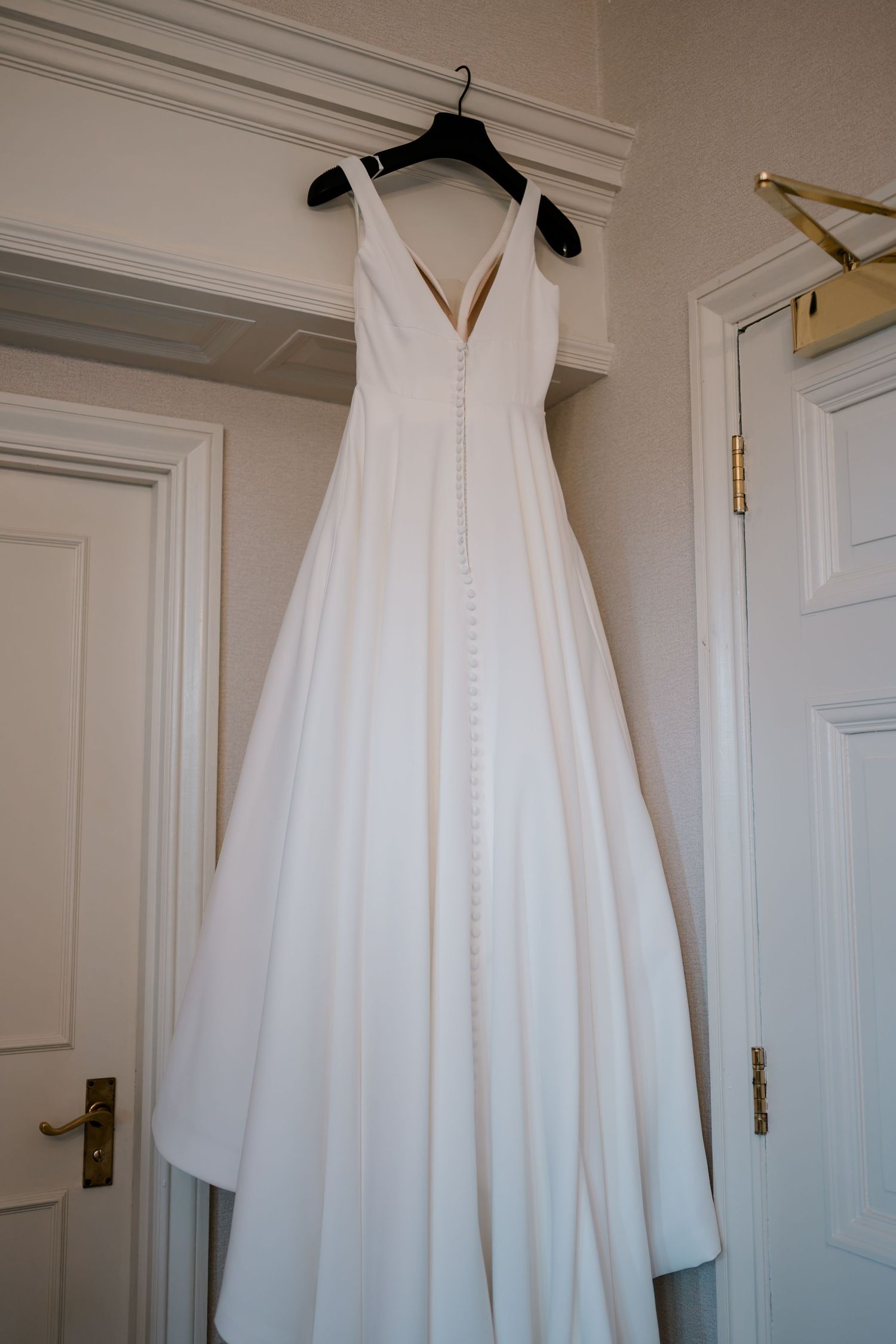 White V-neck wedding dress with a full skirt and covered button back hanging on a black hanger from a door frame.