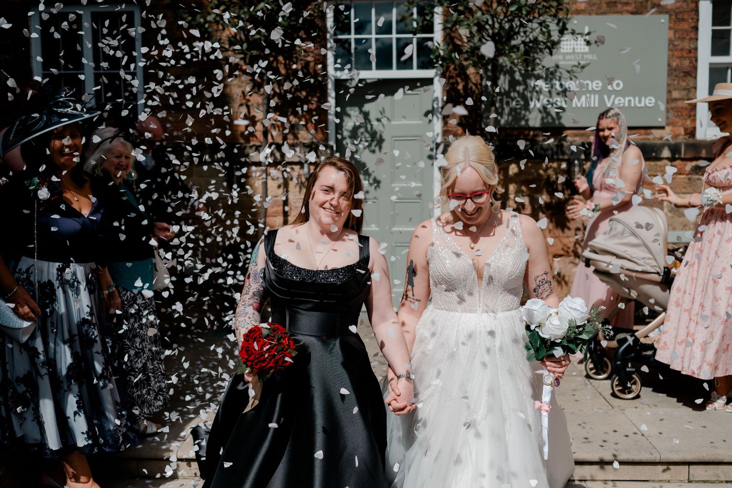 Two brides hold hands walking through confetti shower on their wedding day, one in black gown, one in white lace dress.