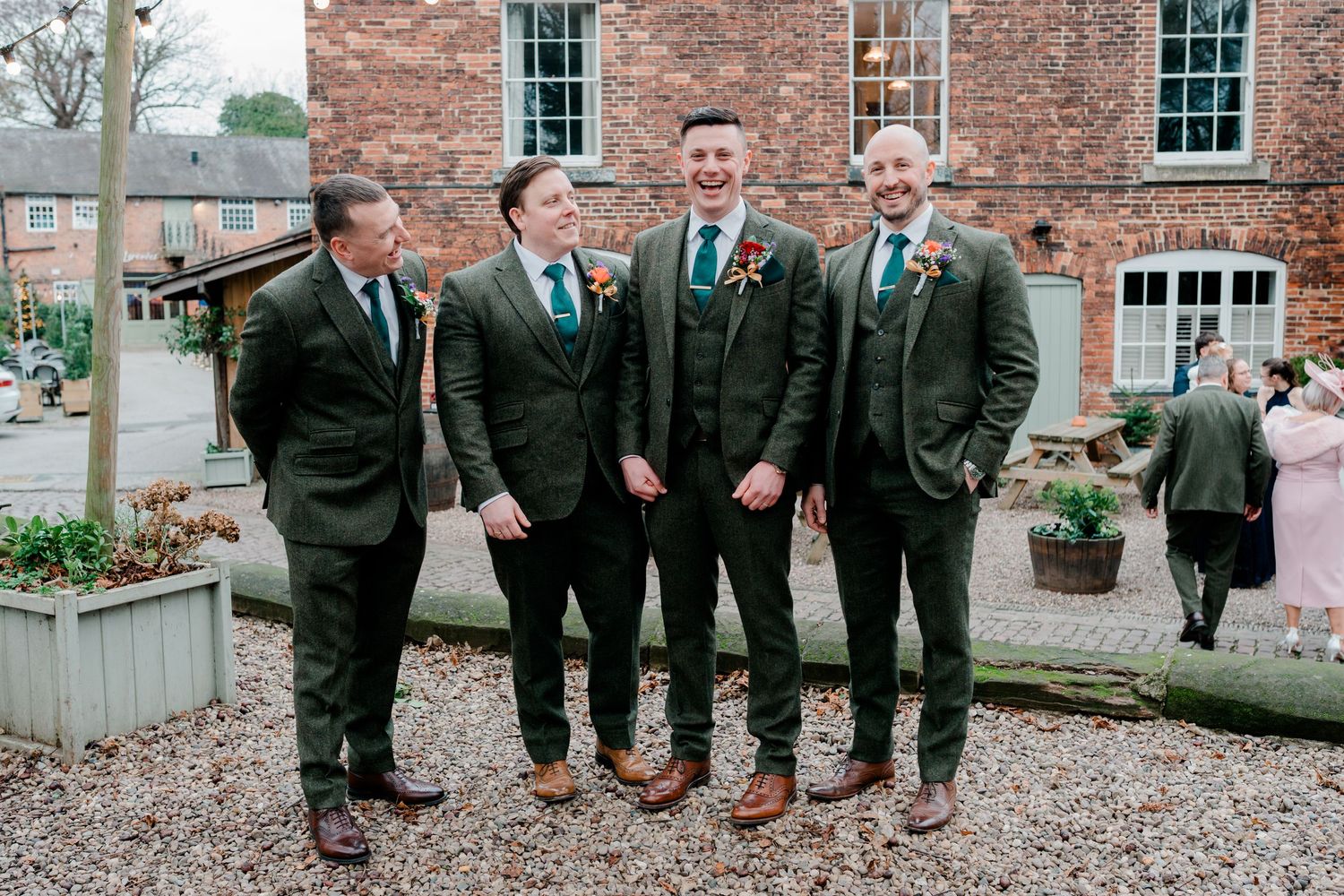 Four groomsmen in green tweed suits laughing together outside a brick wedding venue.