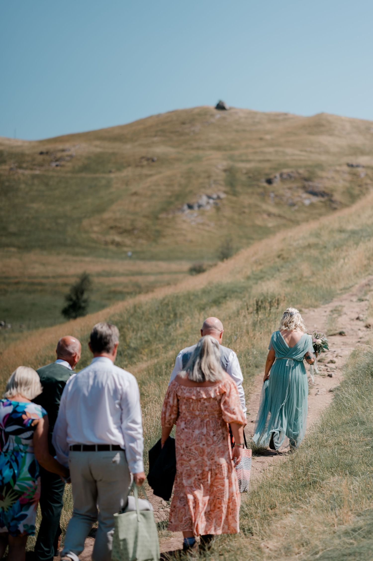 Group of wedding guests walking up a scenic hillside trail on a sunny day, with a woman in a teal gown leading the way.