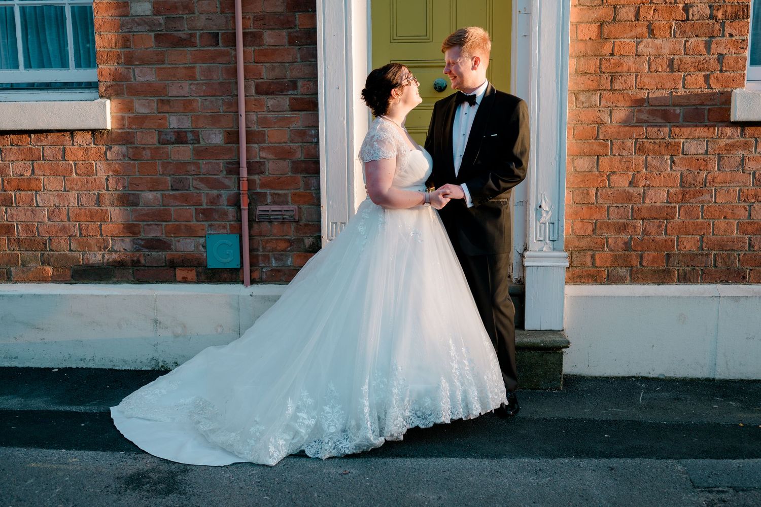 Bride in white ballgown and groom in black tuxedo share a romantic moment outside a brick building on their wedding day.