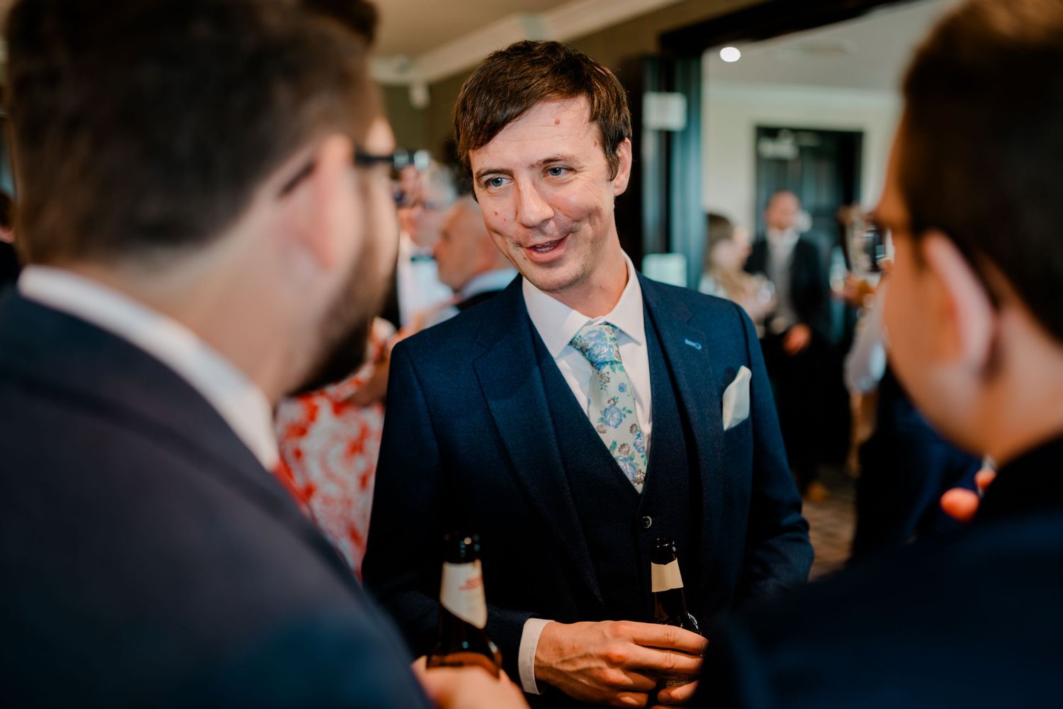 A guest in a navy suit and floral tie socializes at a formal event while holding a drink.