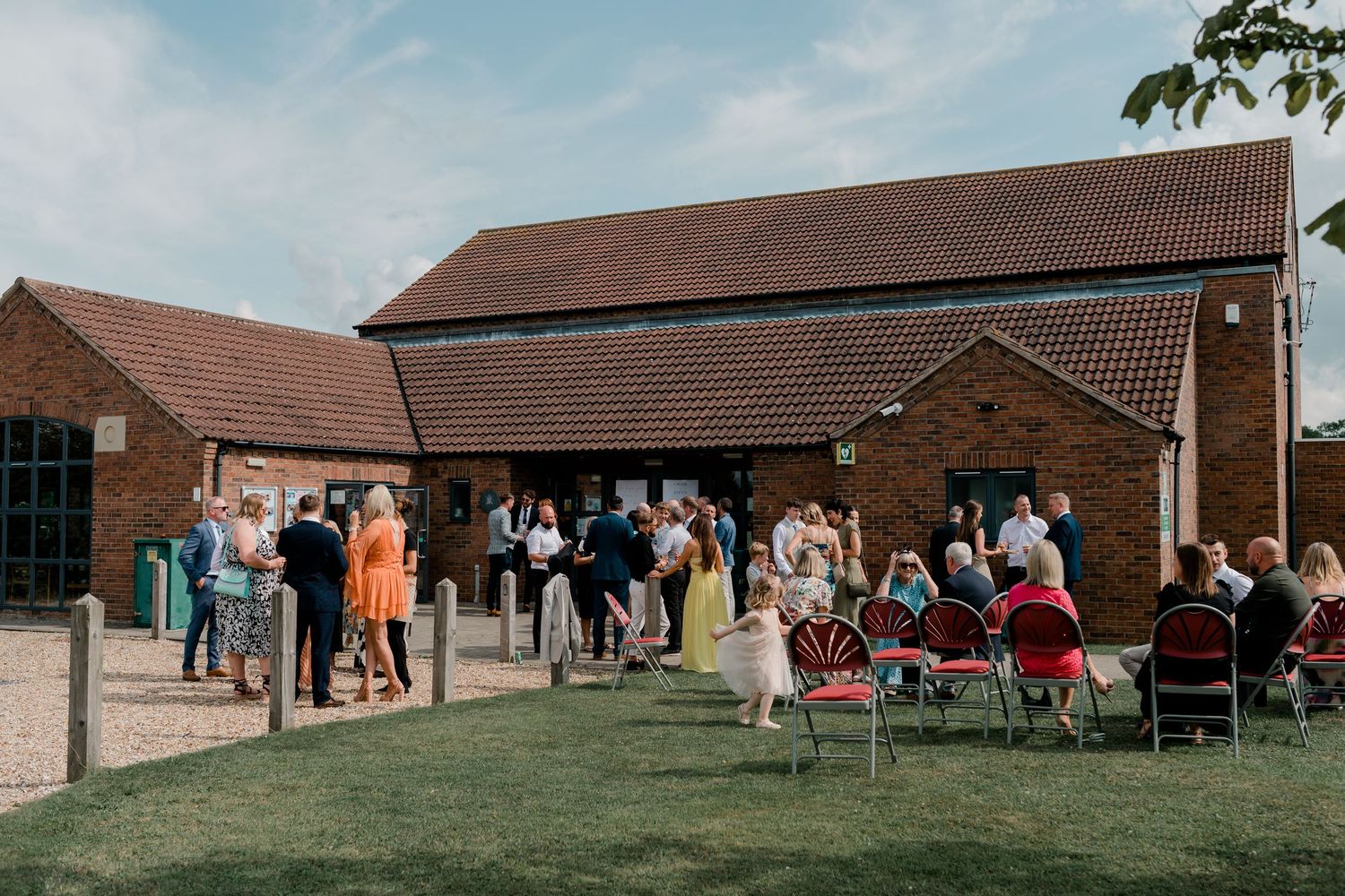 Wedding guests gather outside a brick building during a sunny afternoon reception.