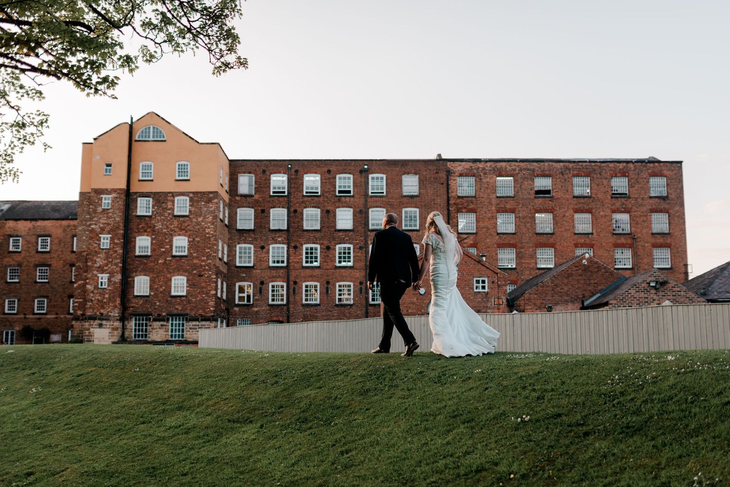 Elegant bride and groom walk hand in hand past a historic red brick building at dusk.