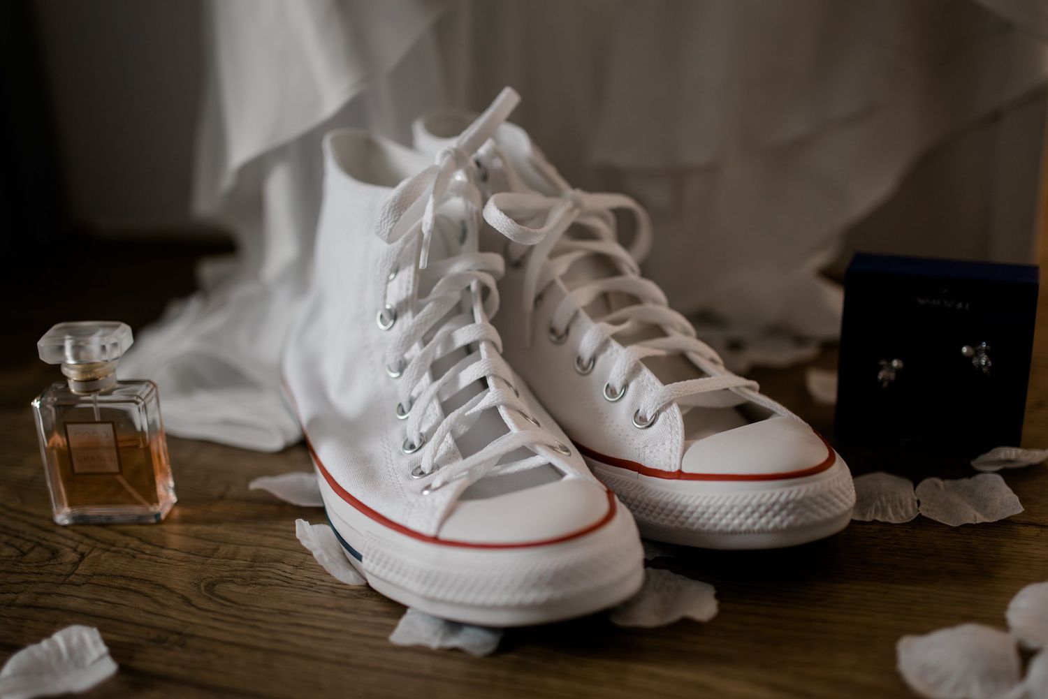 White Converse high-top sneakers styled with wedding accessories including perfume and earrings on a wooden floor.