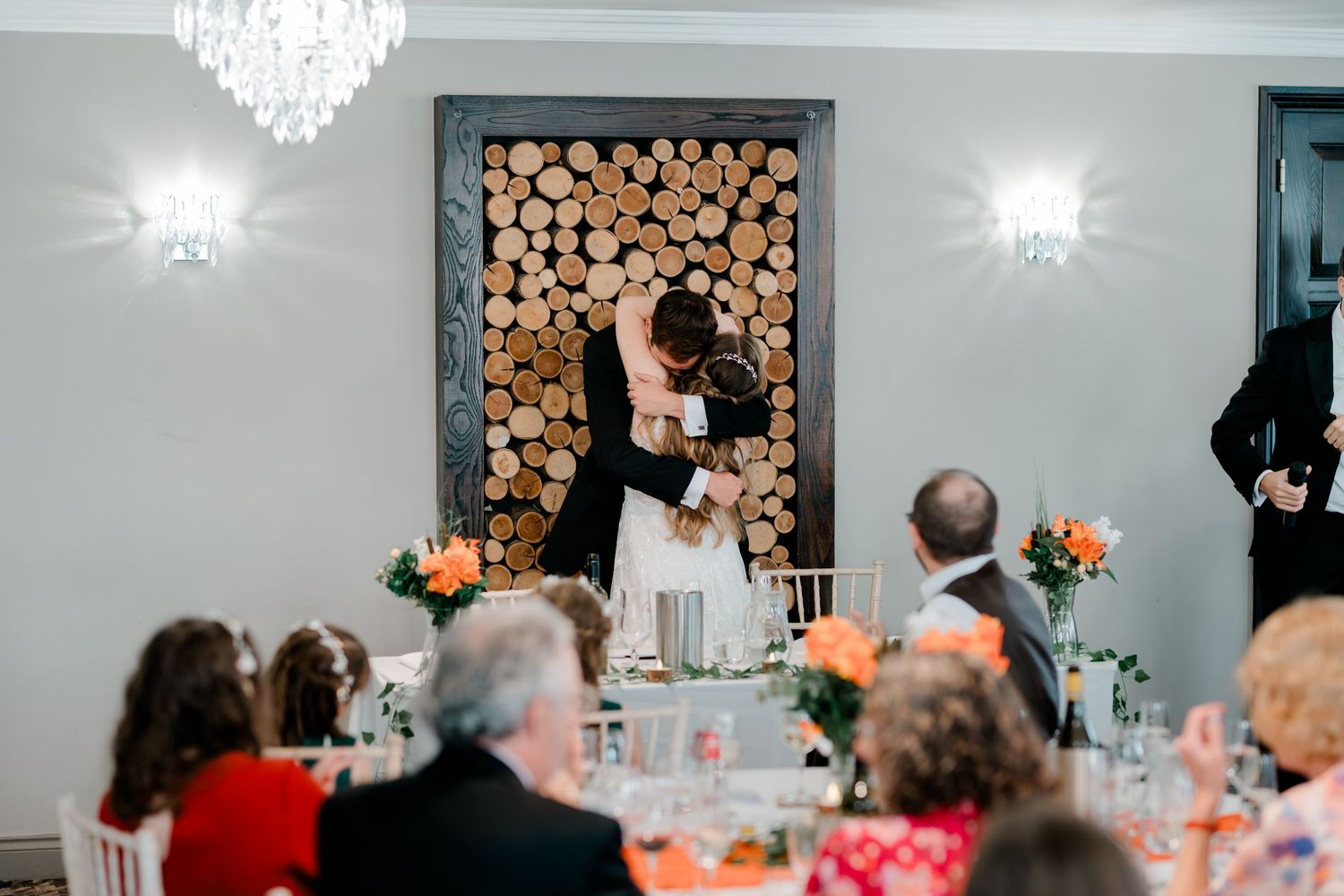 Bride and groom share a romantic kiss at wedding reception with decorative log wall feature behind them.