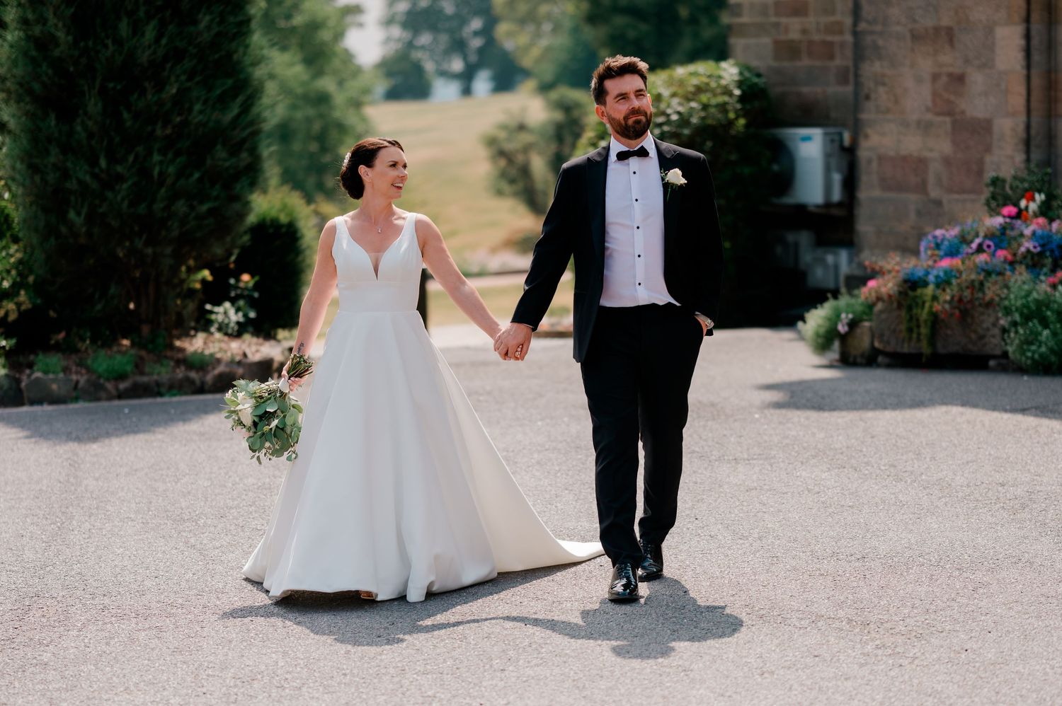 A bride in a white gown holding a bouquet walks hand in hand with a groom in a black suit and bow tie along a sunlit garden path.