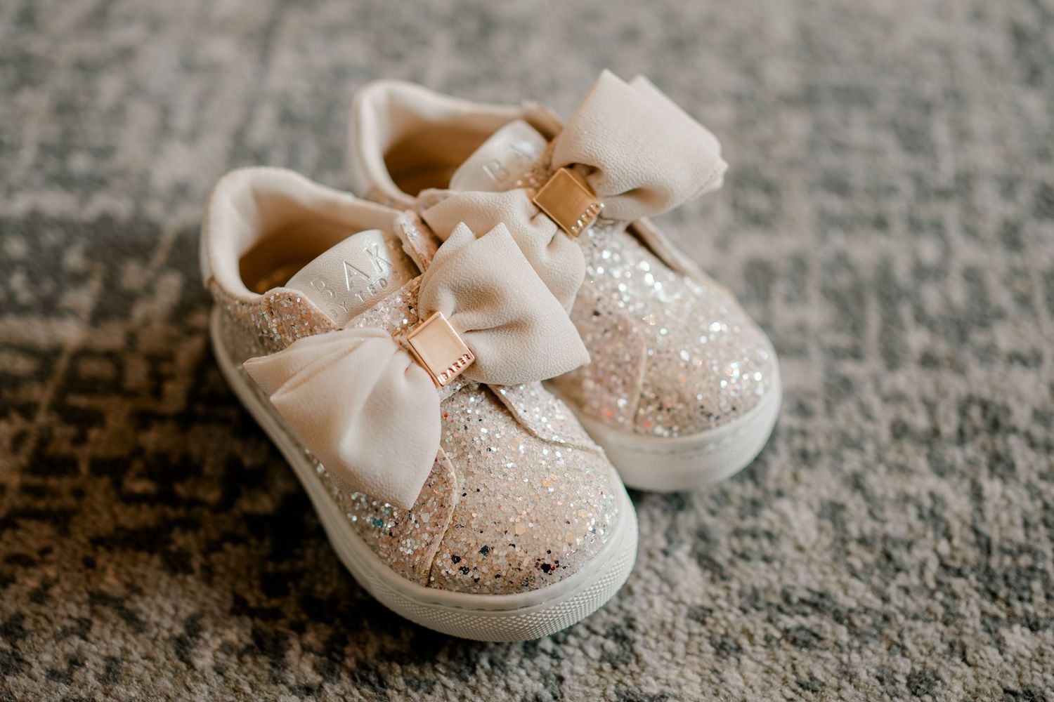 Pair of glittery gold toddler sneakers with cream satin bows and gold buckles on a gray textured rug.
