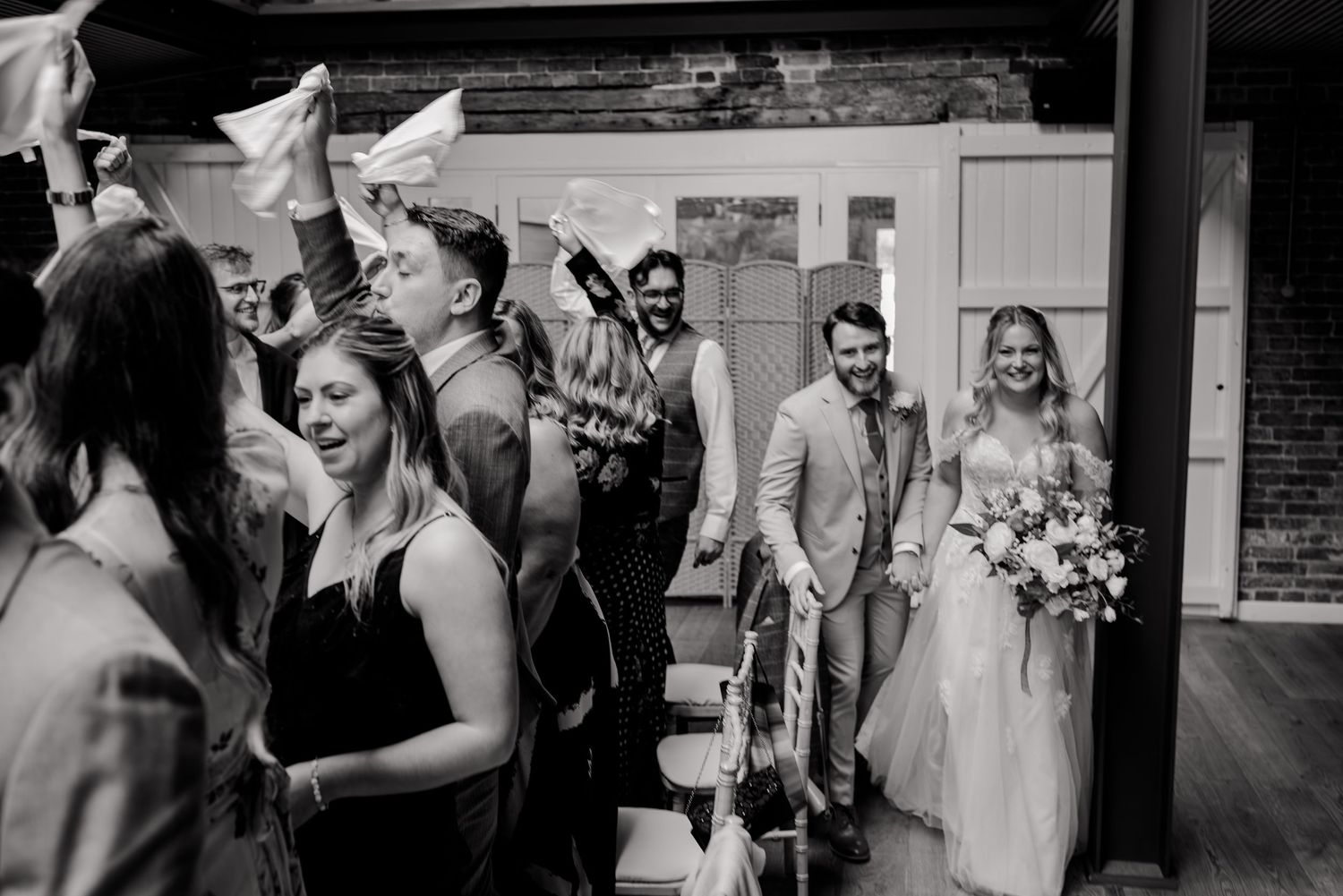 Bride and groom make grand entrance at wedding reception as guests celebrate with raised hands.