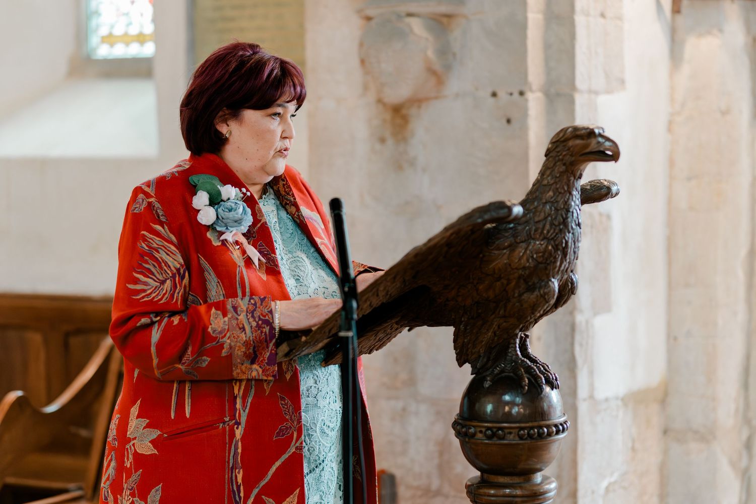Woman in red floral coat reading at an eagle lectern during a church wedding ceremony.