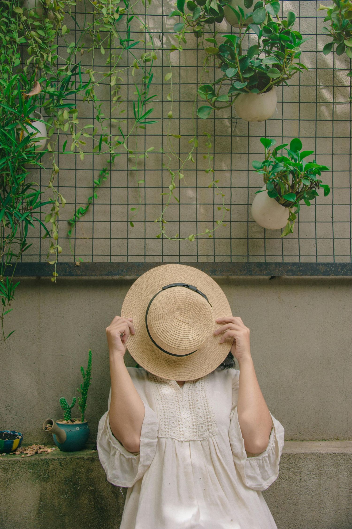 A person in white dress holds a straw hat in front of their face against a plant-covered wall.