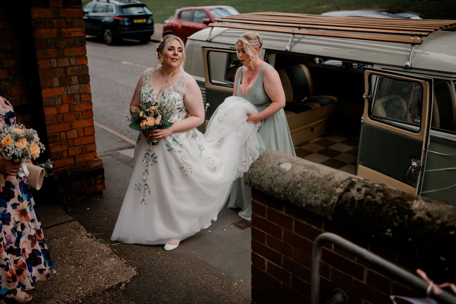 Wedding party gathered outside brick building with vintage cars in background.