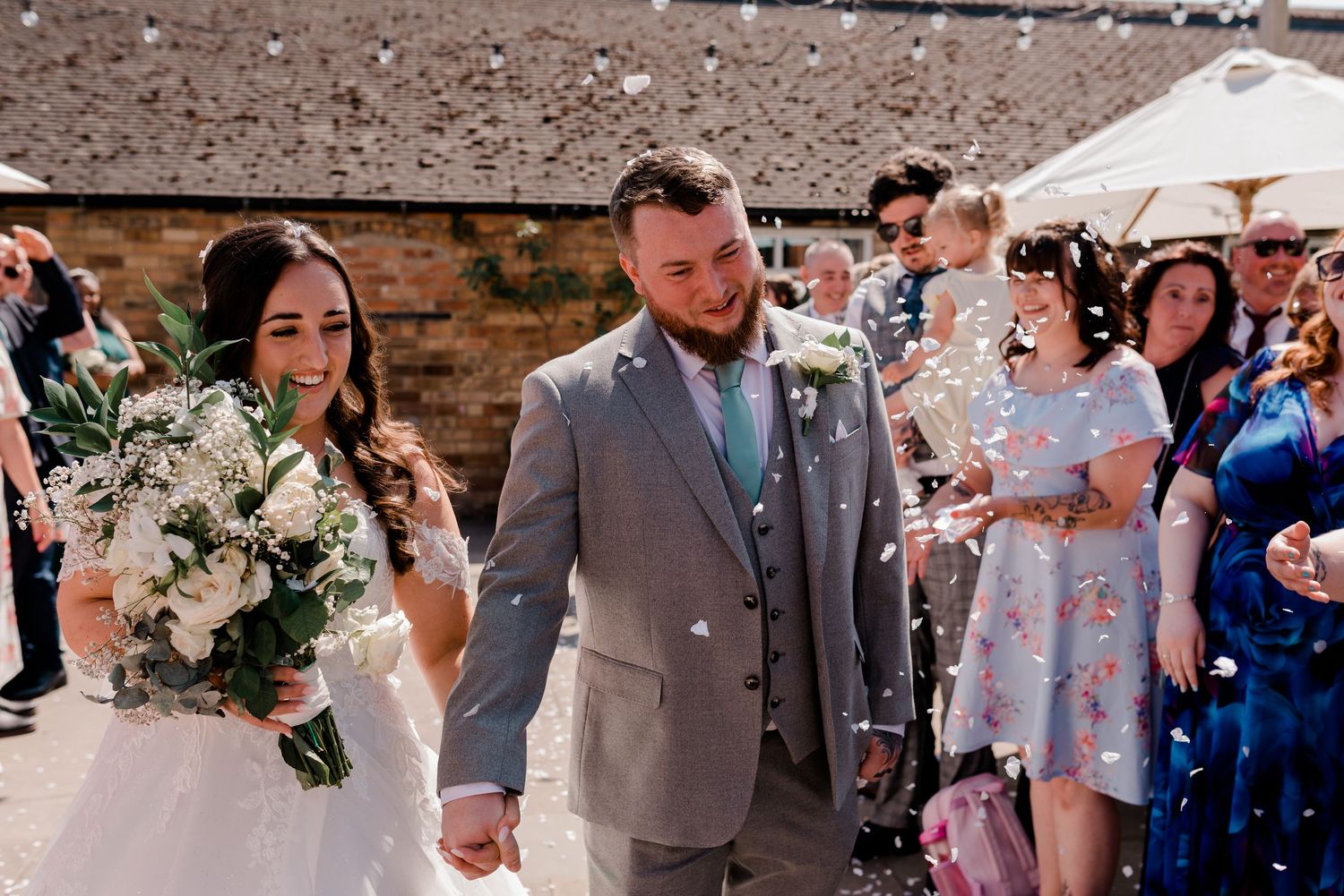 A wedding couple walks together past guests at an outdoor ceremony with a grey suited groom and white floral bouquet.