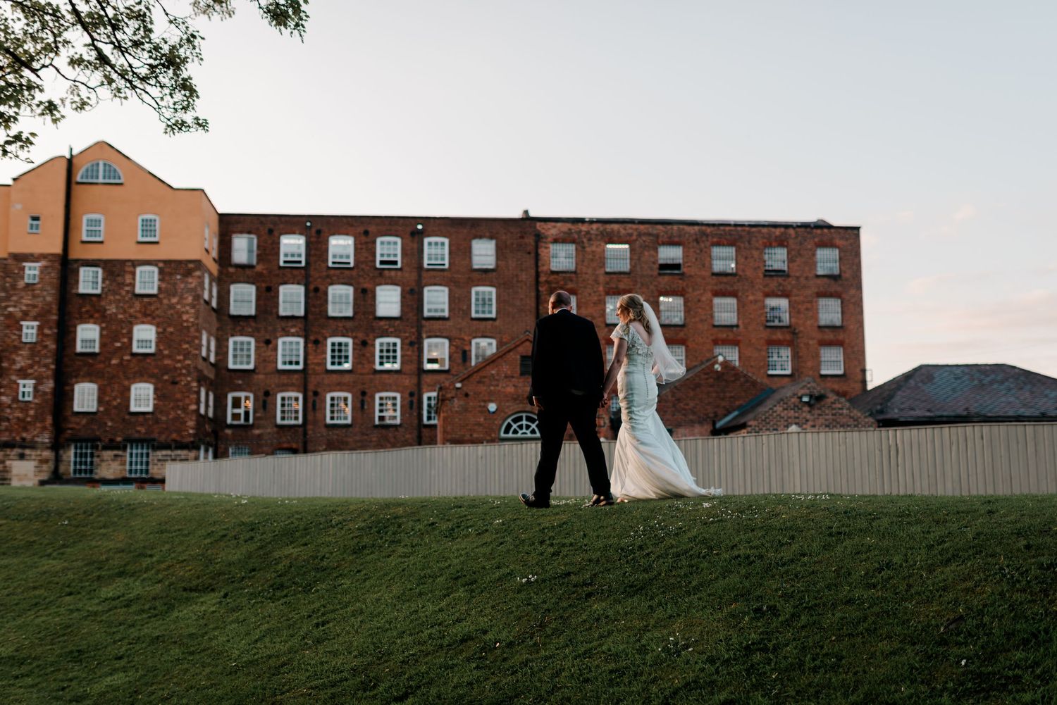 A bride and groom walk hand in hand on green grass before a large historic red brick building at dusk.